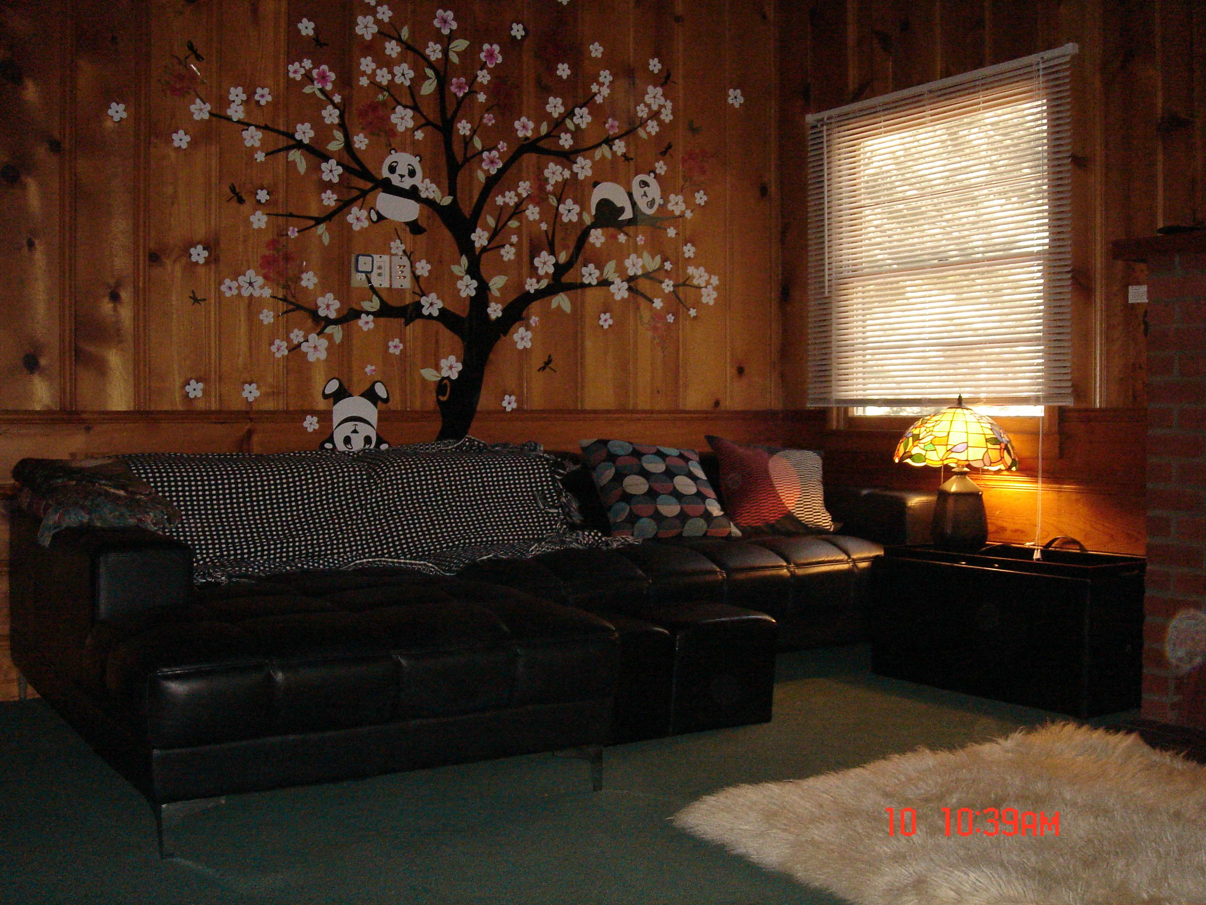 A living room with a black sectional sofa, a decorative tree mural on the wood-paneled wall, and a lamp beside a window.