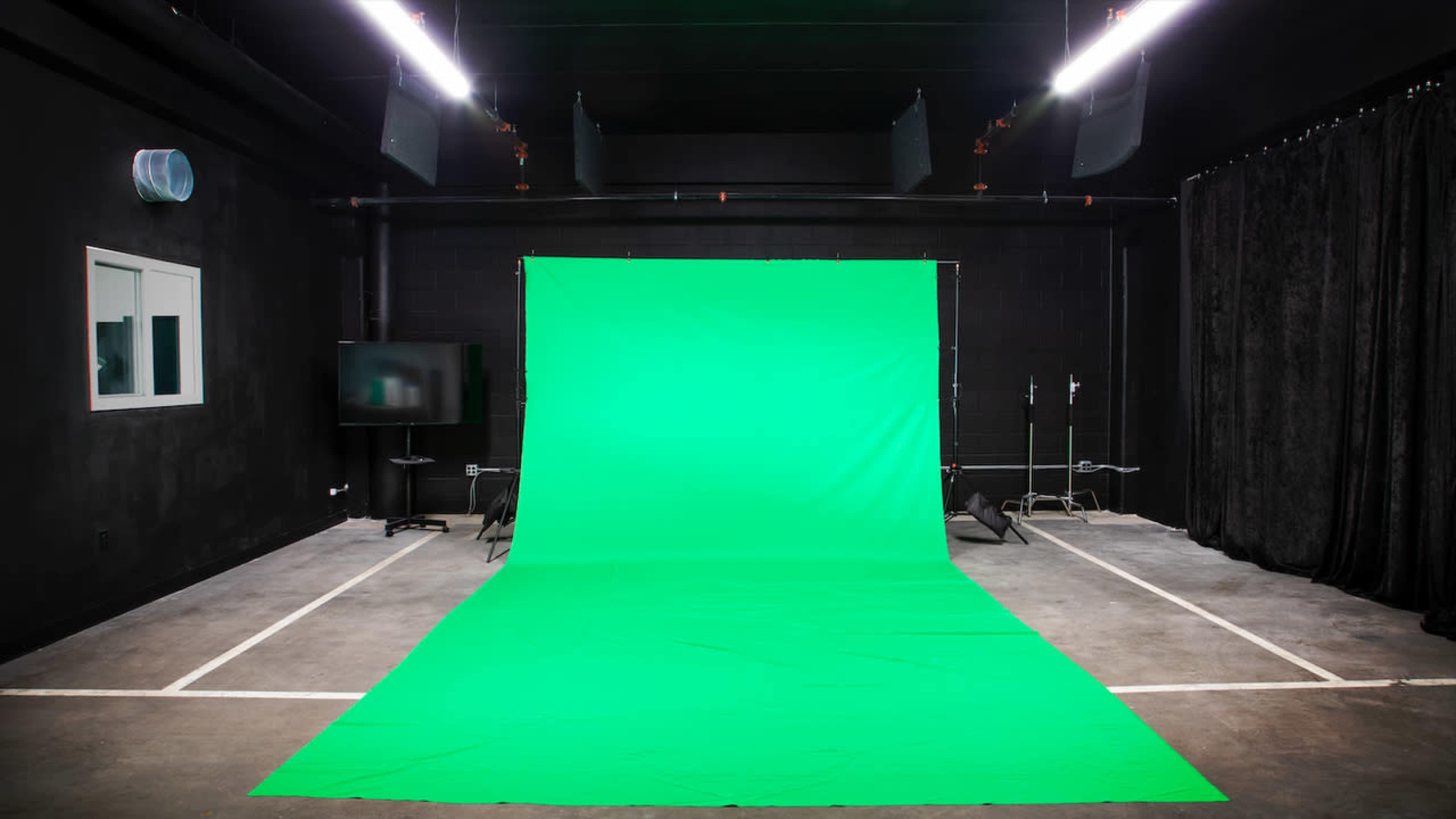 An empty studio space with a large green screen set up against a black wall, illuminated by overhead lights.