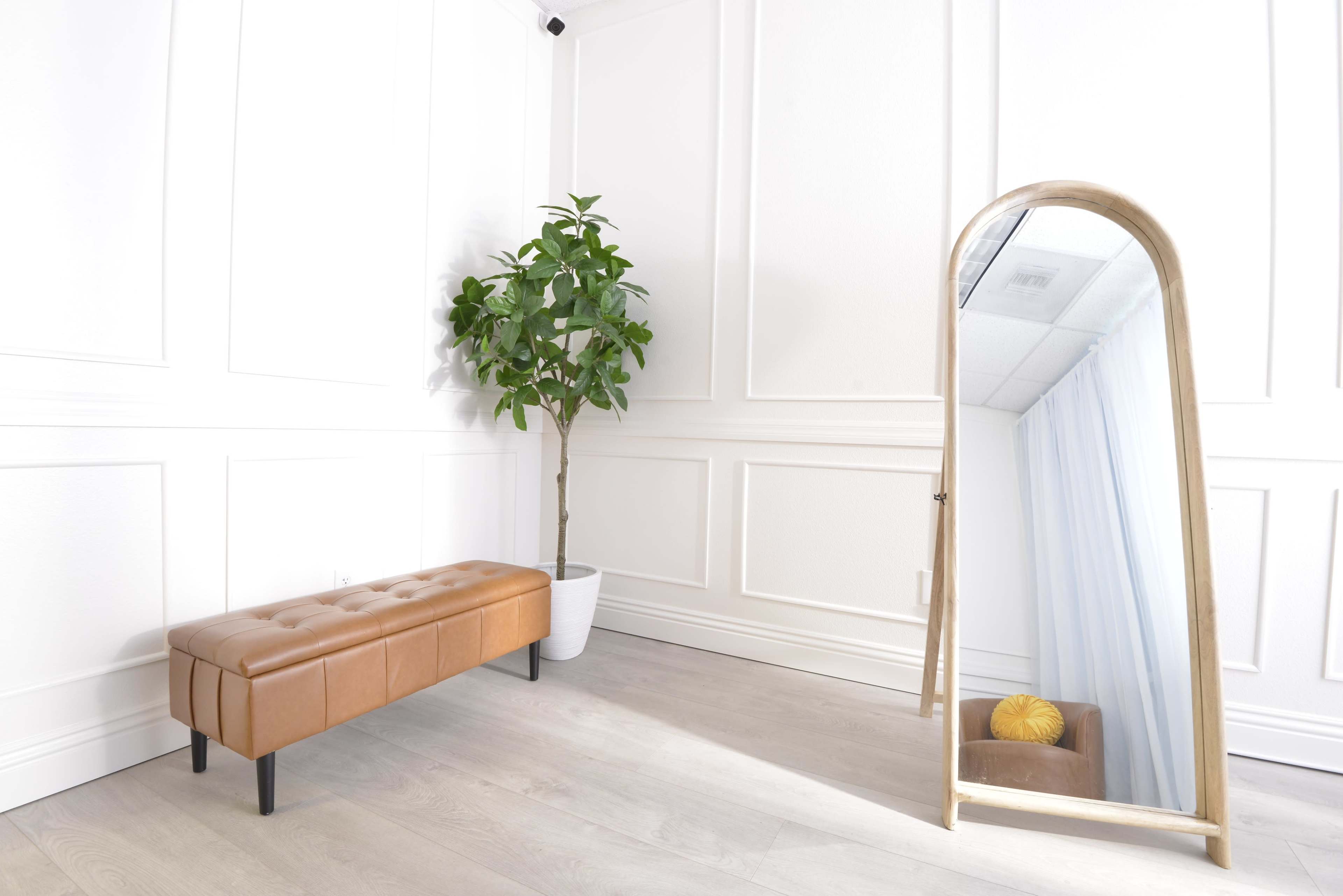 The image shows a minimalist interior with a brown upholstered bench, a potted plant in a white container, and a full-length mirror against the wall.