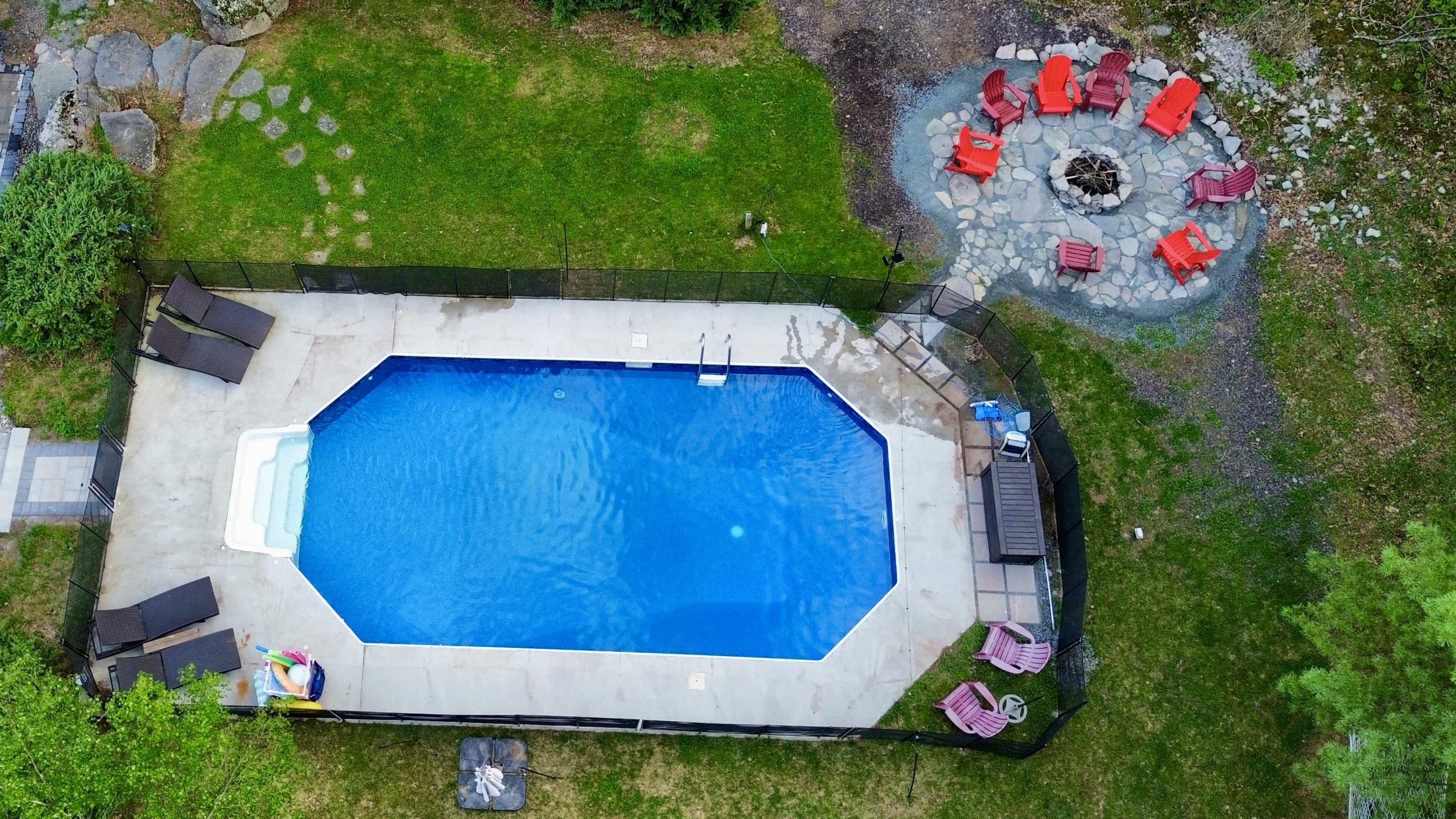 The image shows an aerial view of a rectangular swimming pool surrounded by a fenced area, with a circular fire pit and red chairs arranged nearby on a grassy lawn.