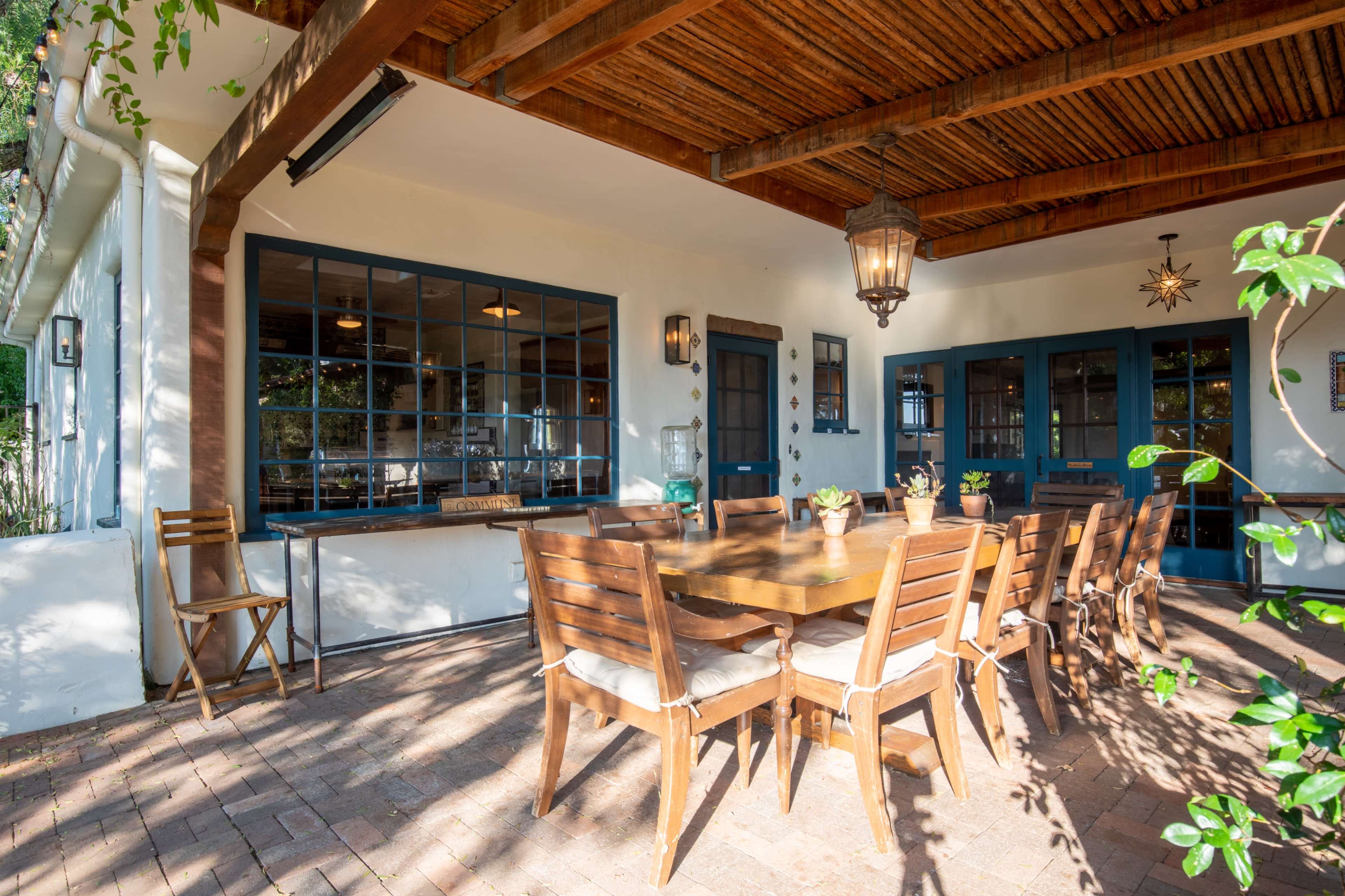 The image shows a spacious outdoor dining area with a large wooden table and chairs beneath a wooden-beamed roof.