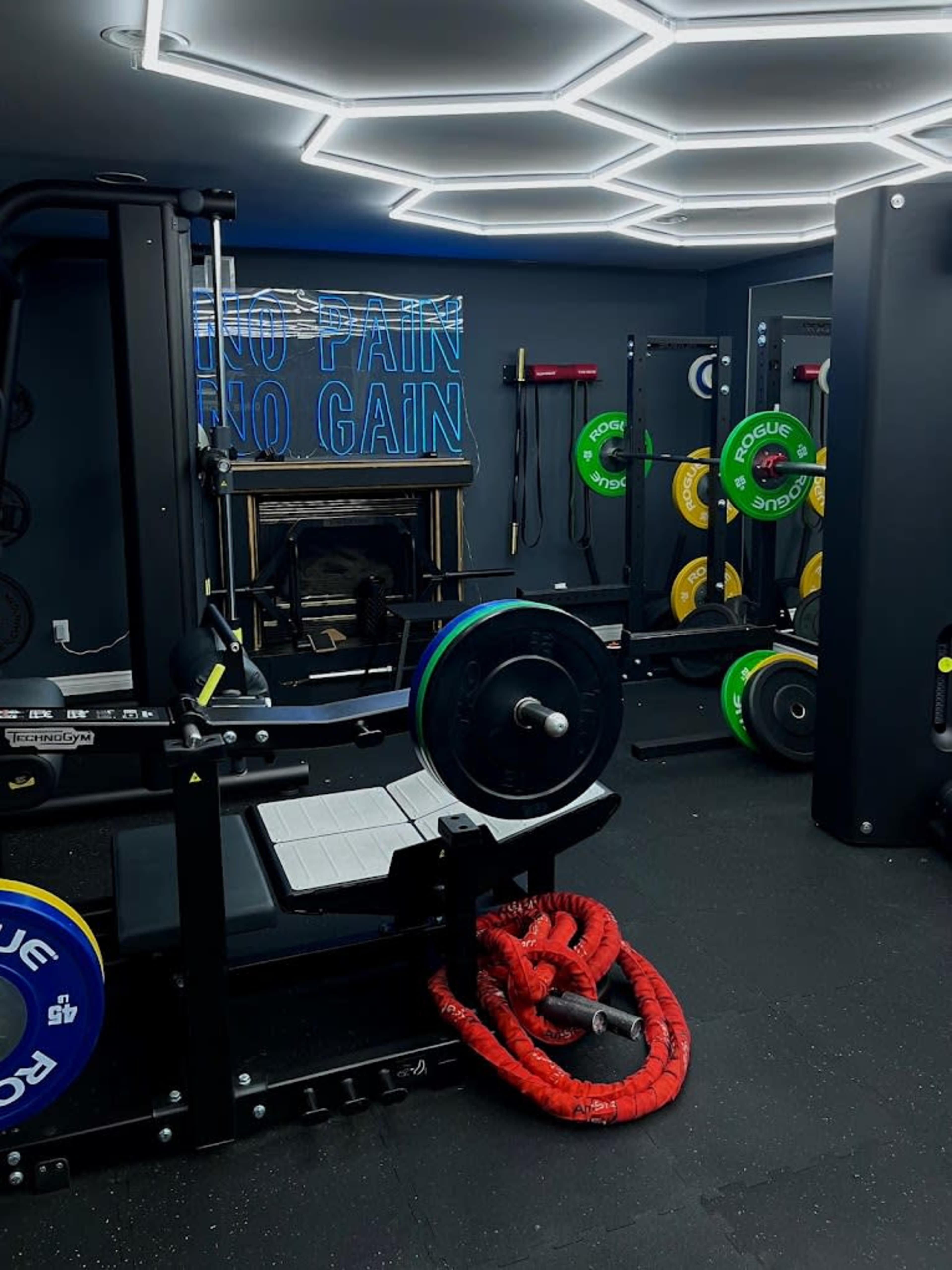 A modern gym room with various weightlifting equipment, including a bench press and colorful weight plates, along with red ropes on the floor and a motivational sign on the wall.