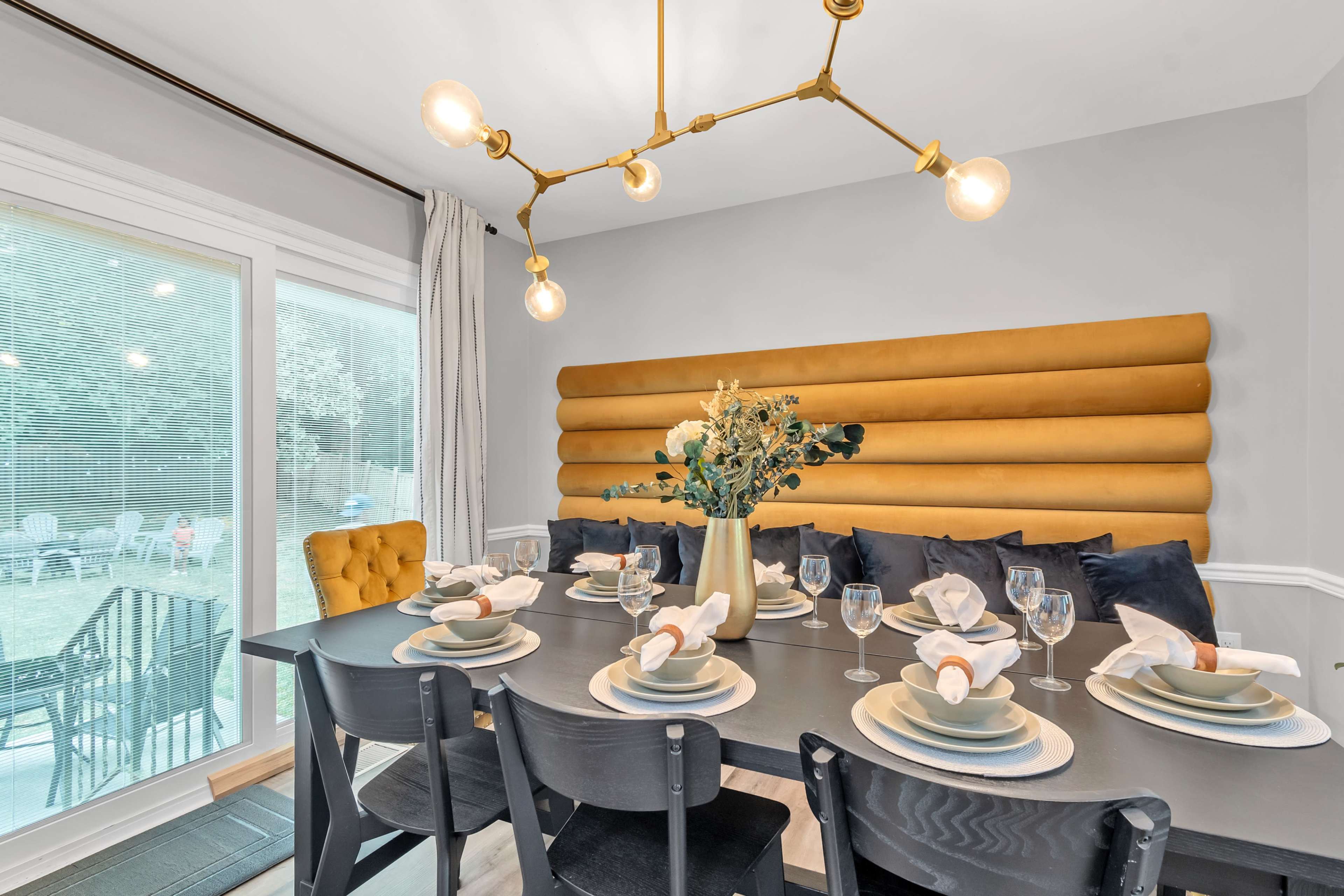 A dining room features a long table set with plates, glasses, and decorative touches, surrounded by a yellow upholstered wall and a modern light fixture.