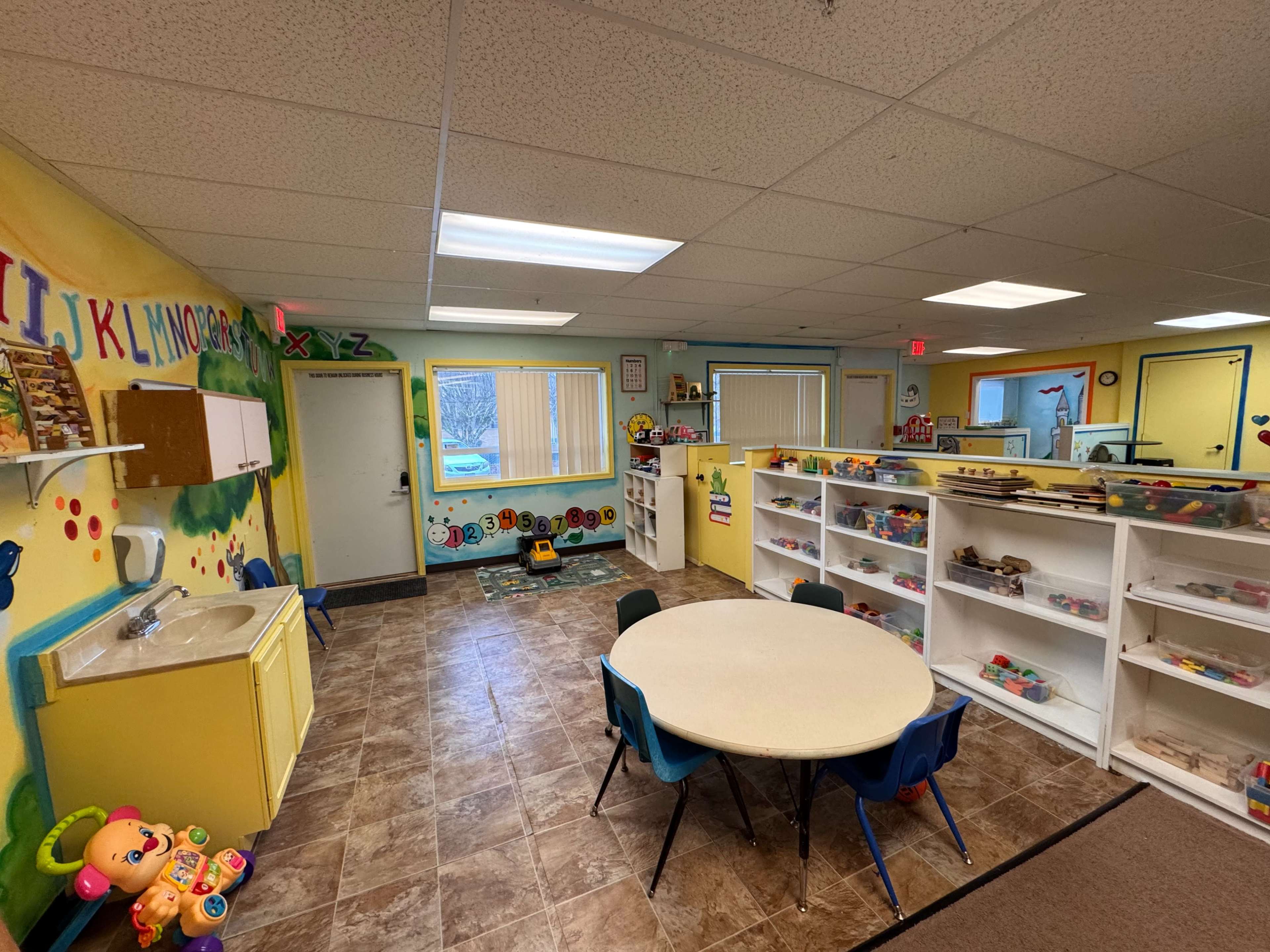 The image shows a brightly colored classroom for young children, featuring playful decorations, a circular table with chairs, and shelves filled with toys and educational materials.