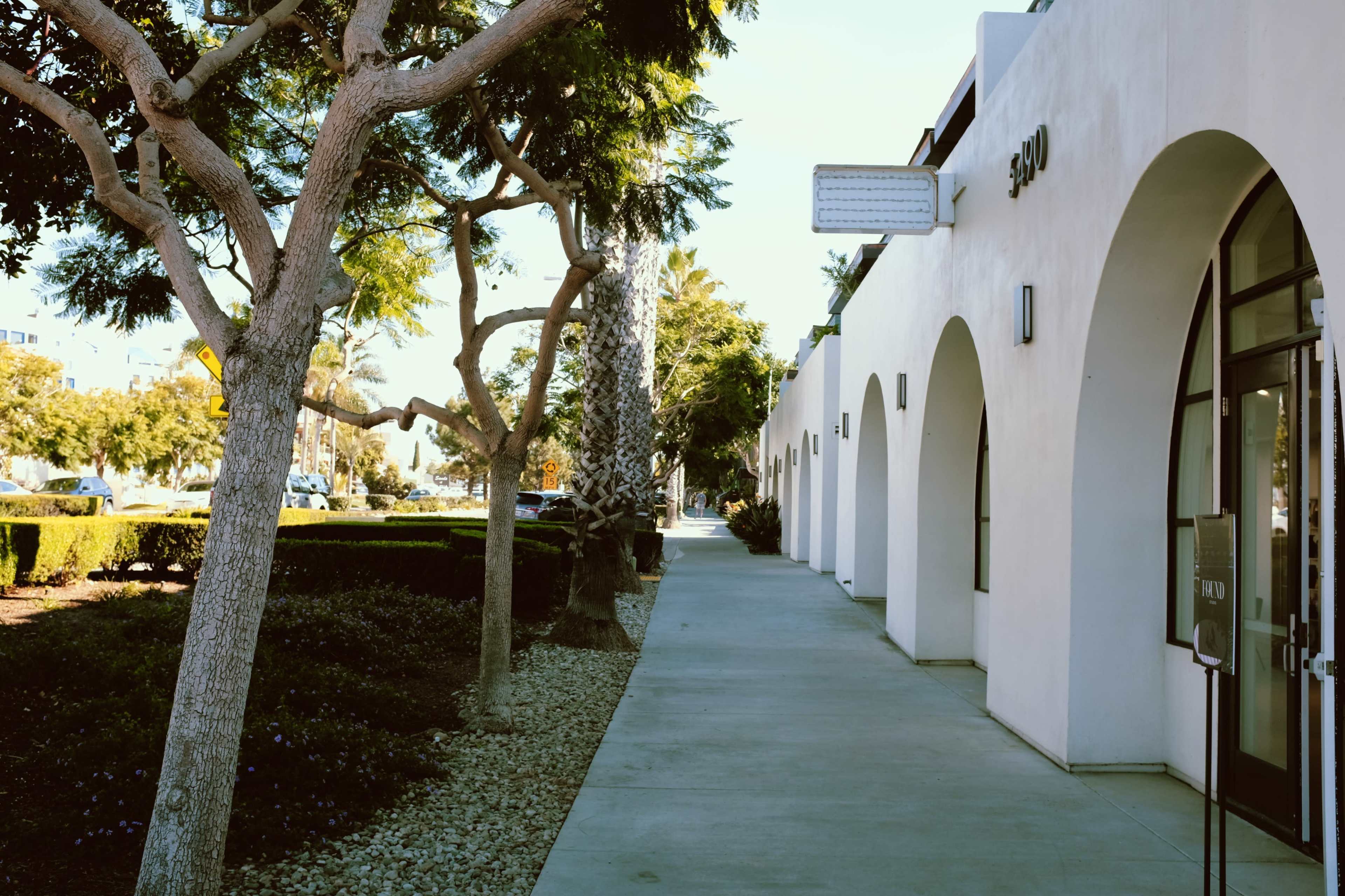 A pedestrian sidewalk runs alongside a row of white buildings, flanked by trees and neatly trimmed hedges.