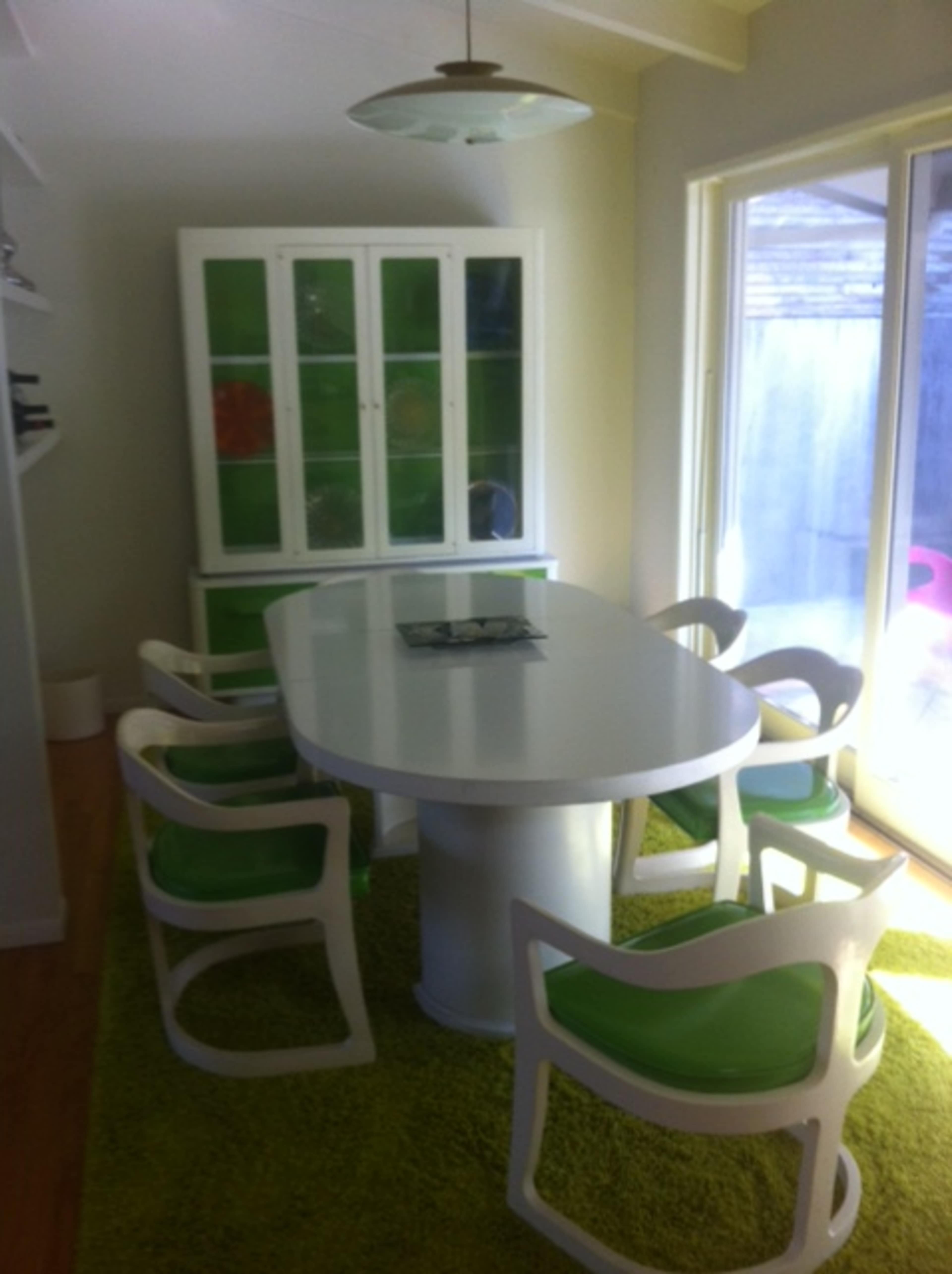 A white oval dining table surrounded by six green and white chairs sits on a green rug in a brightly lit dining area with large windows and a glass cabinet.