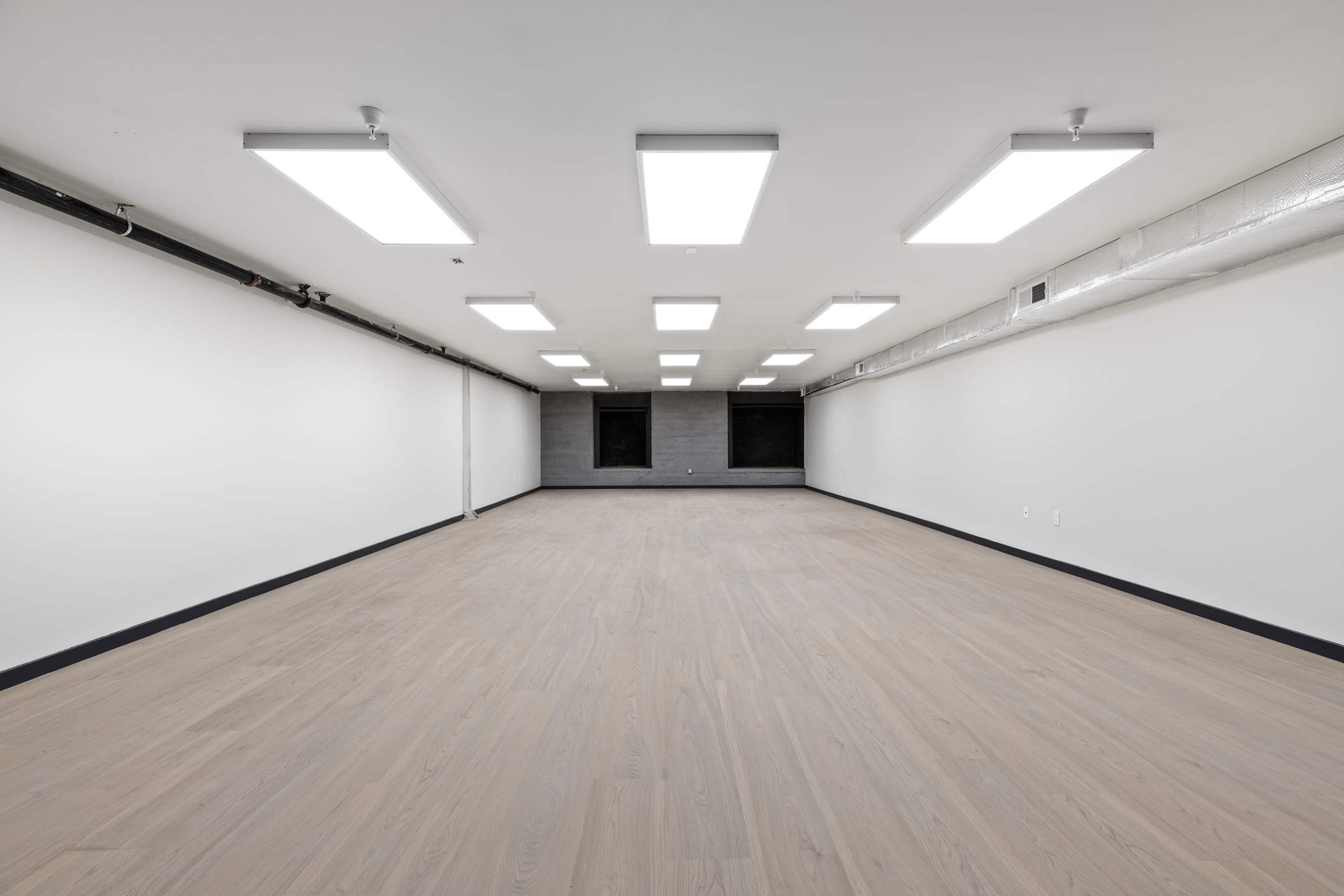 The image shows a spacious, empty room with polished wooden flooring and bright overhead lighting.