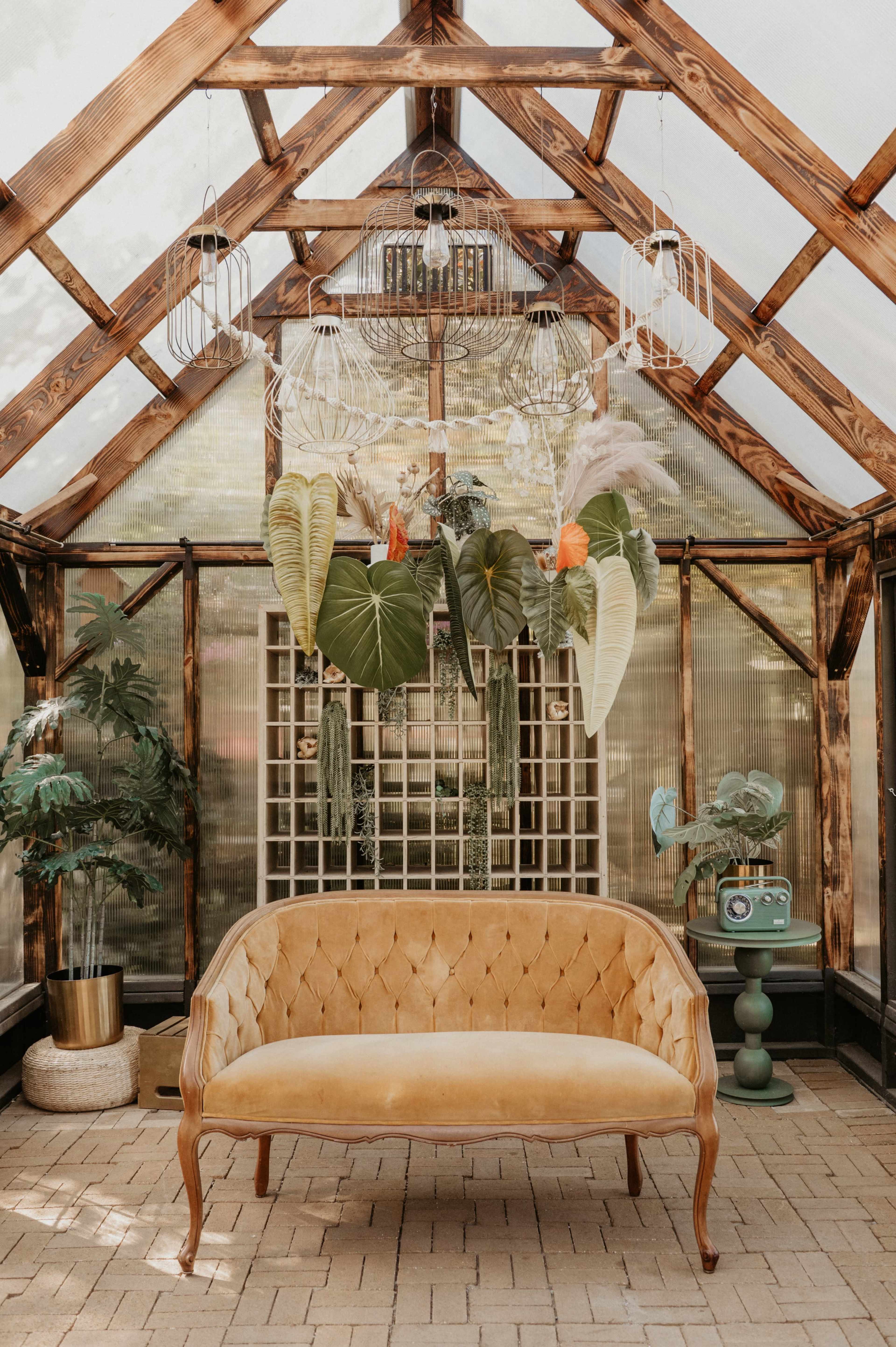 The image depicts a stylish greenhouse interior featuring a tufted yellow sofa, hanging plants, and decorative elements suspended from a wooden frame.
