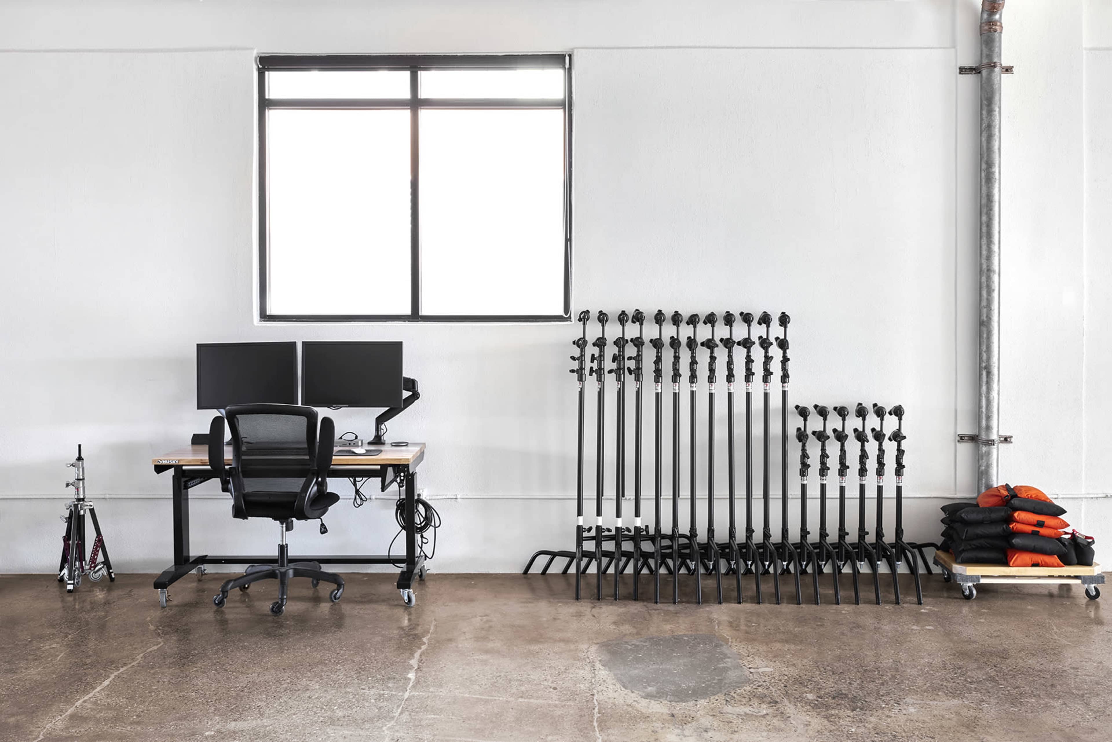 A minimalistic workspace features two computer monitors on a desk, accompanied by a chair and organized equipment stacked neatly against the wall.