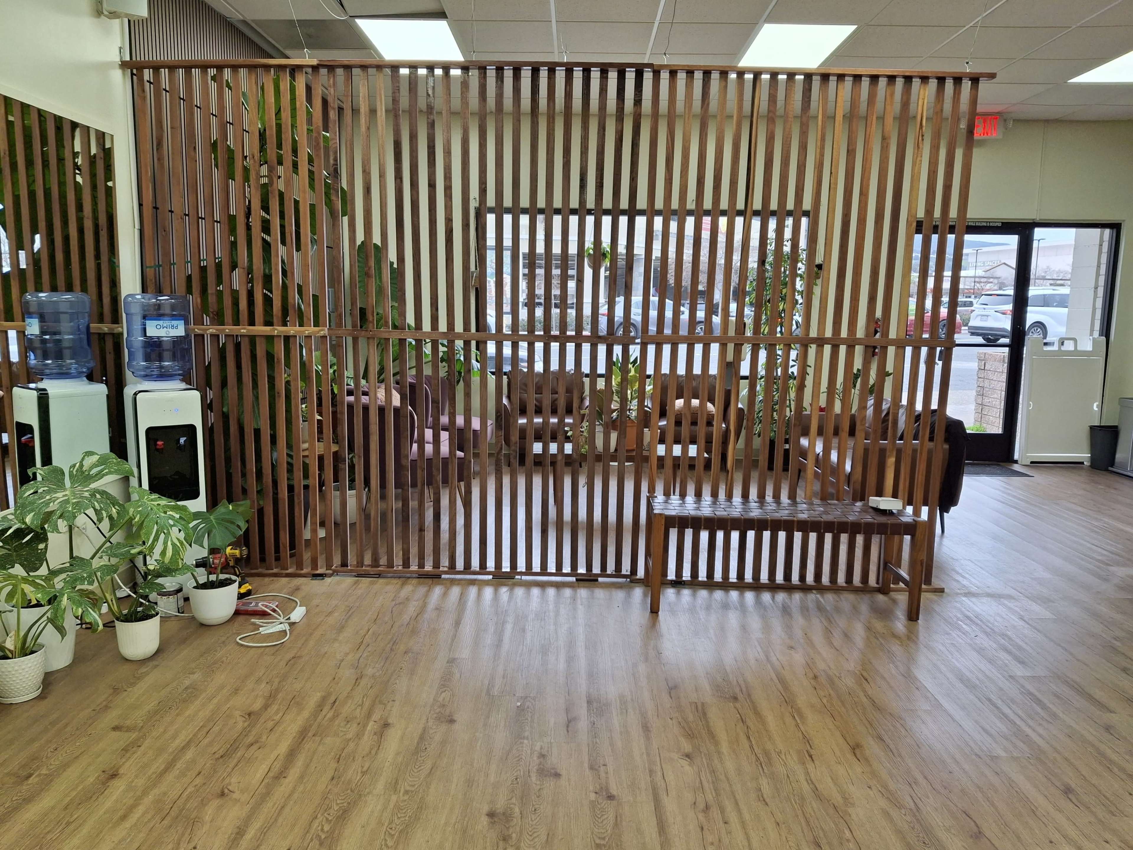 The image shows a spacious indoor setting with a wooden slatted partition dividing a seating area from the entrance, accompanied by water coolers and potted plants.