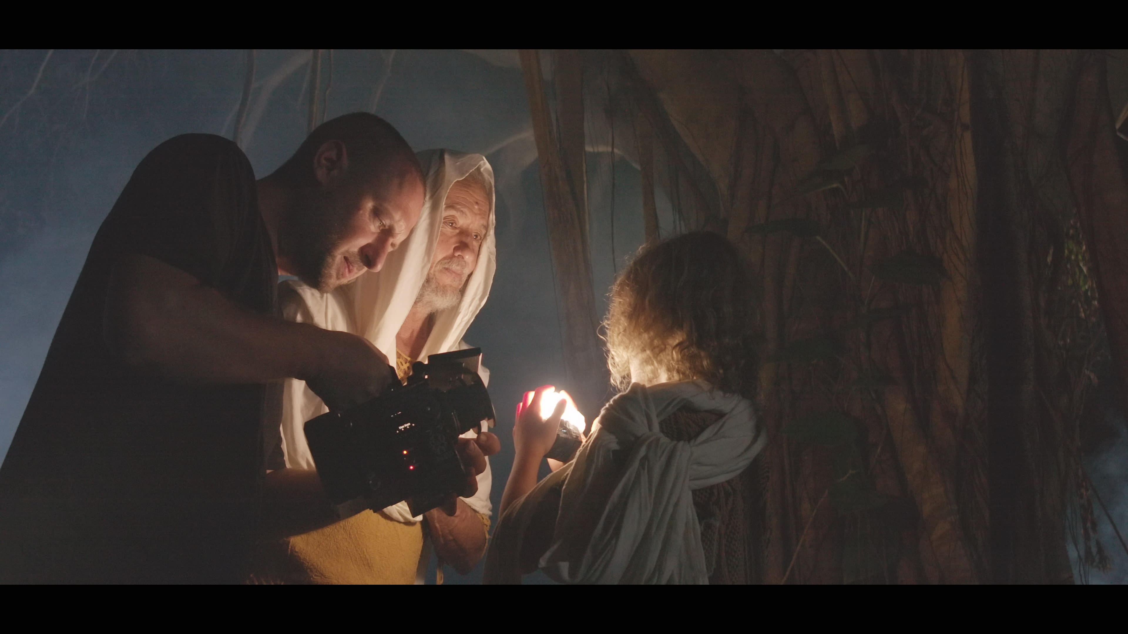 A man with a camera is adjusting equipment while an older man and a child are illuminated by a light in a wooded setting.