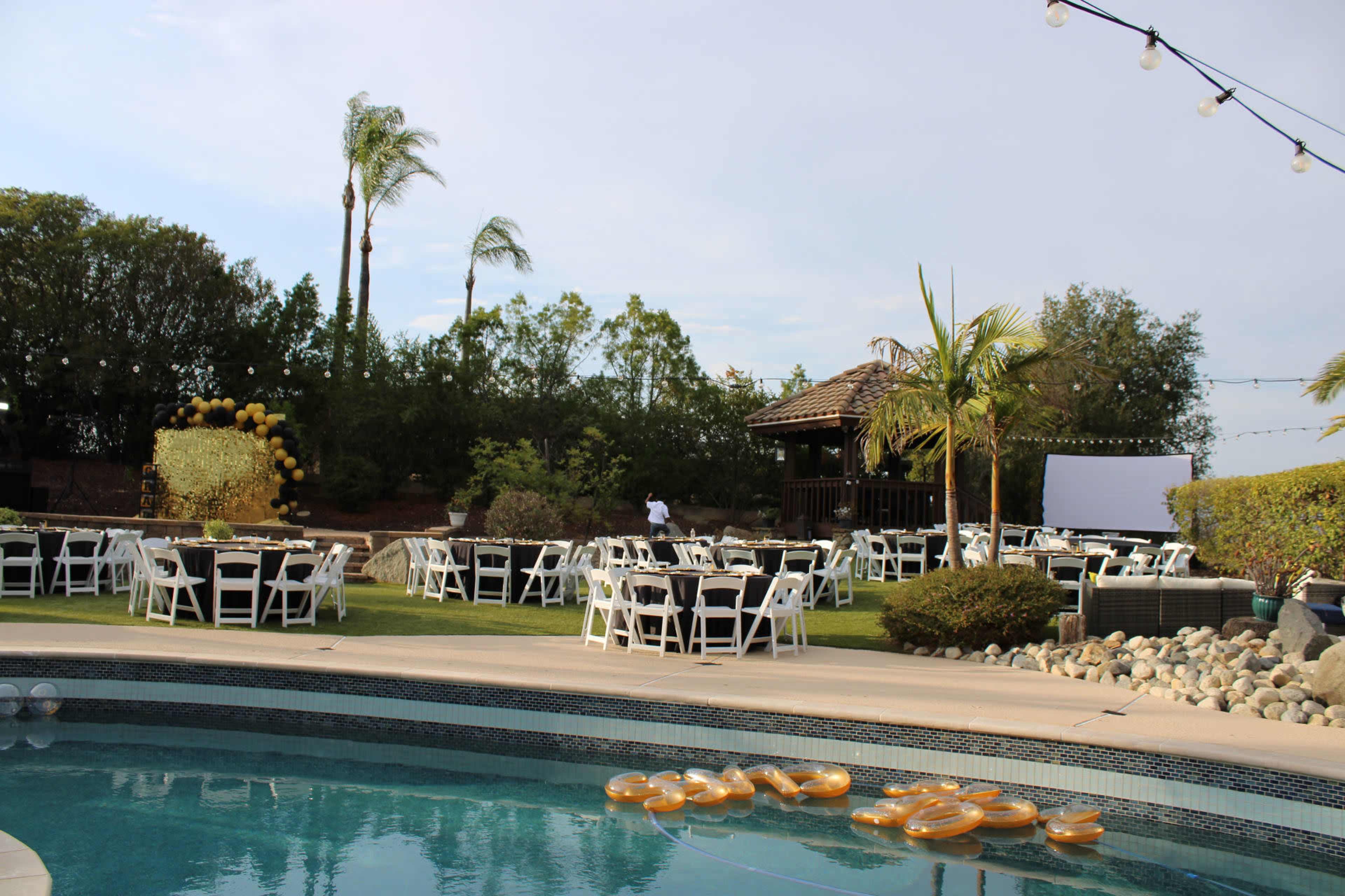 A poolside venue is set with white tables and chairs in a landscaped area, featuring palm trees and a covered pavilion in the background.