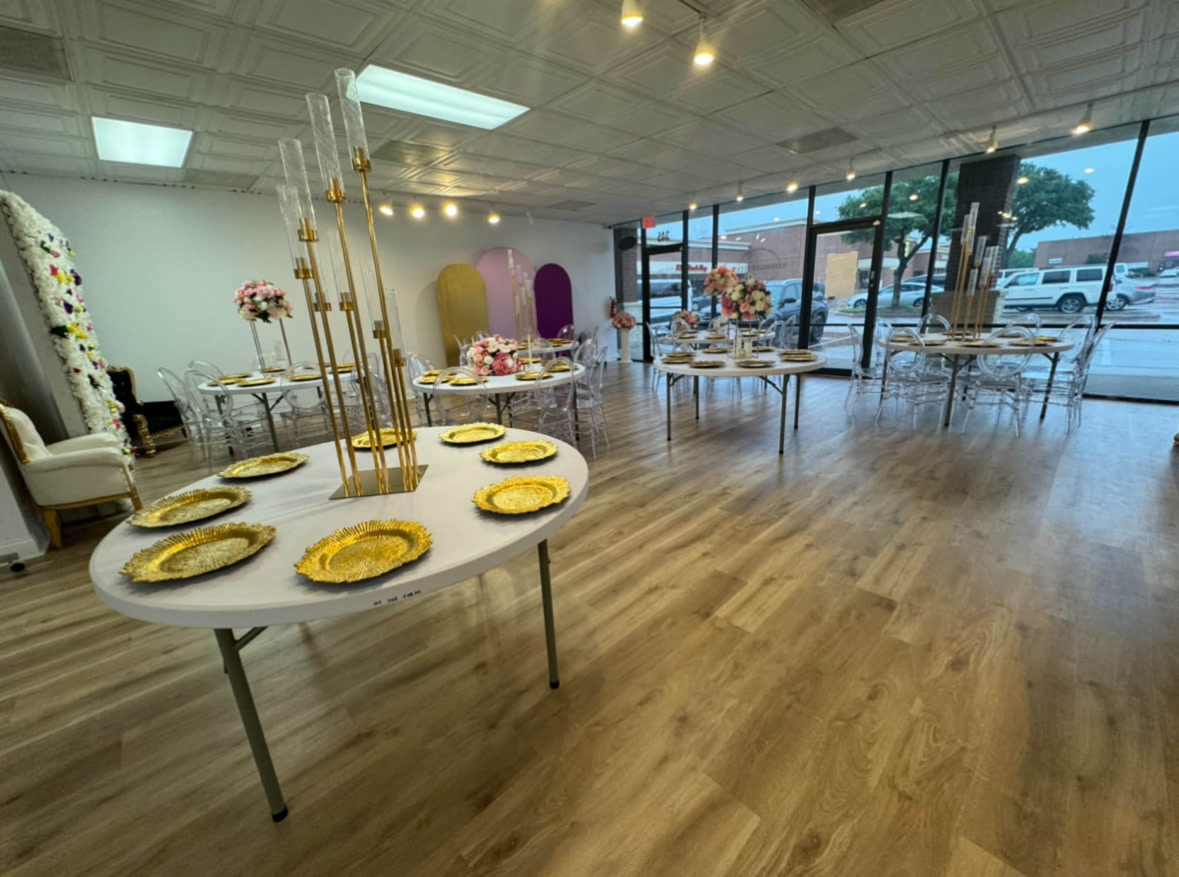A decorated event space with several tables set up, featuring decorative plates and floral arrangements, and large windows allowing natural light to enter.