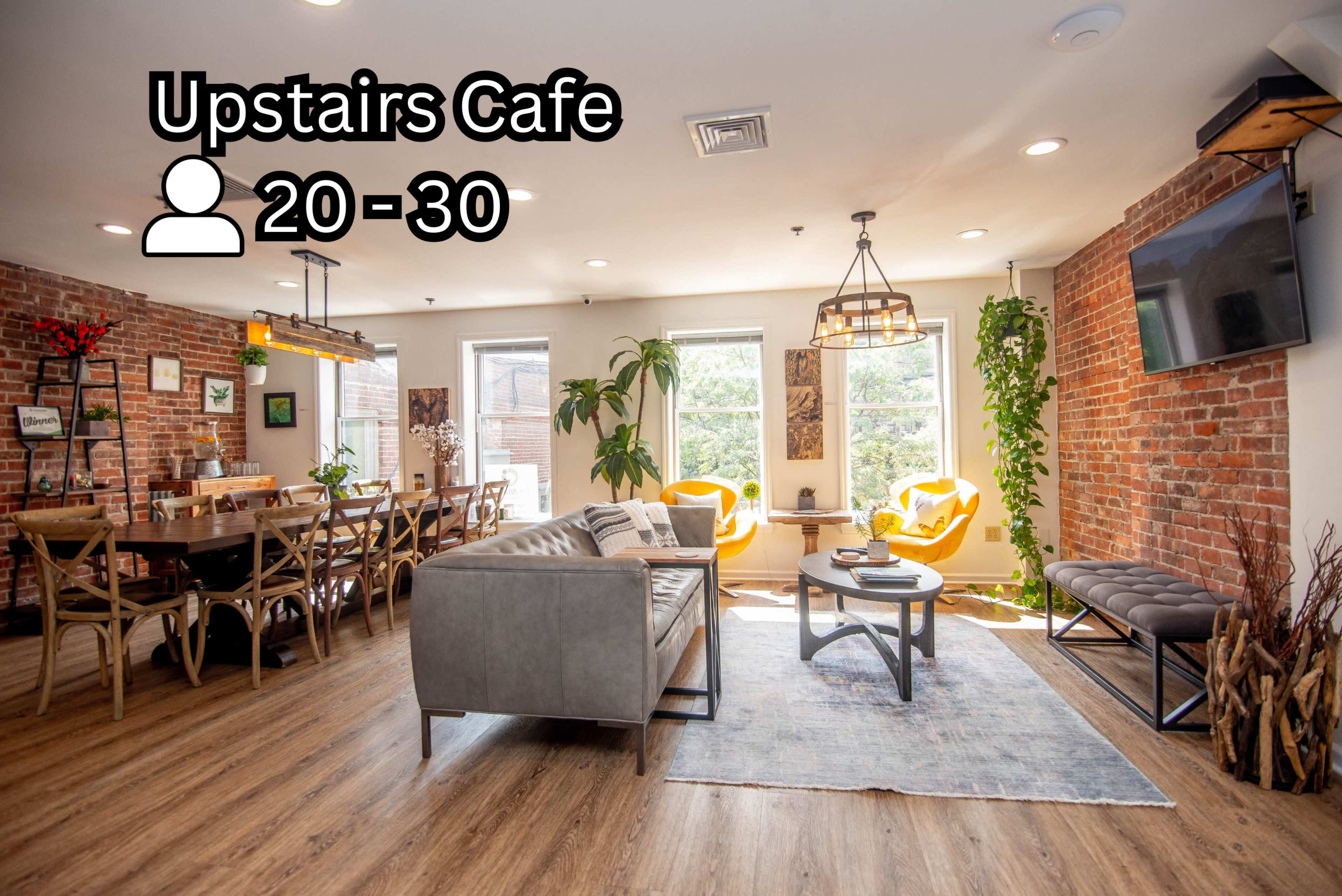 The image shows a cozy cafe with a mix of seating options, including a long dining table and soft armchairs, surrounded by exposed brick walls and large windows.
