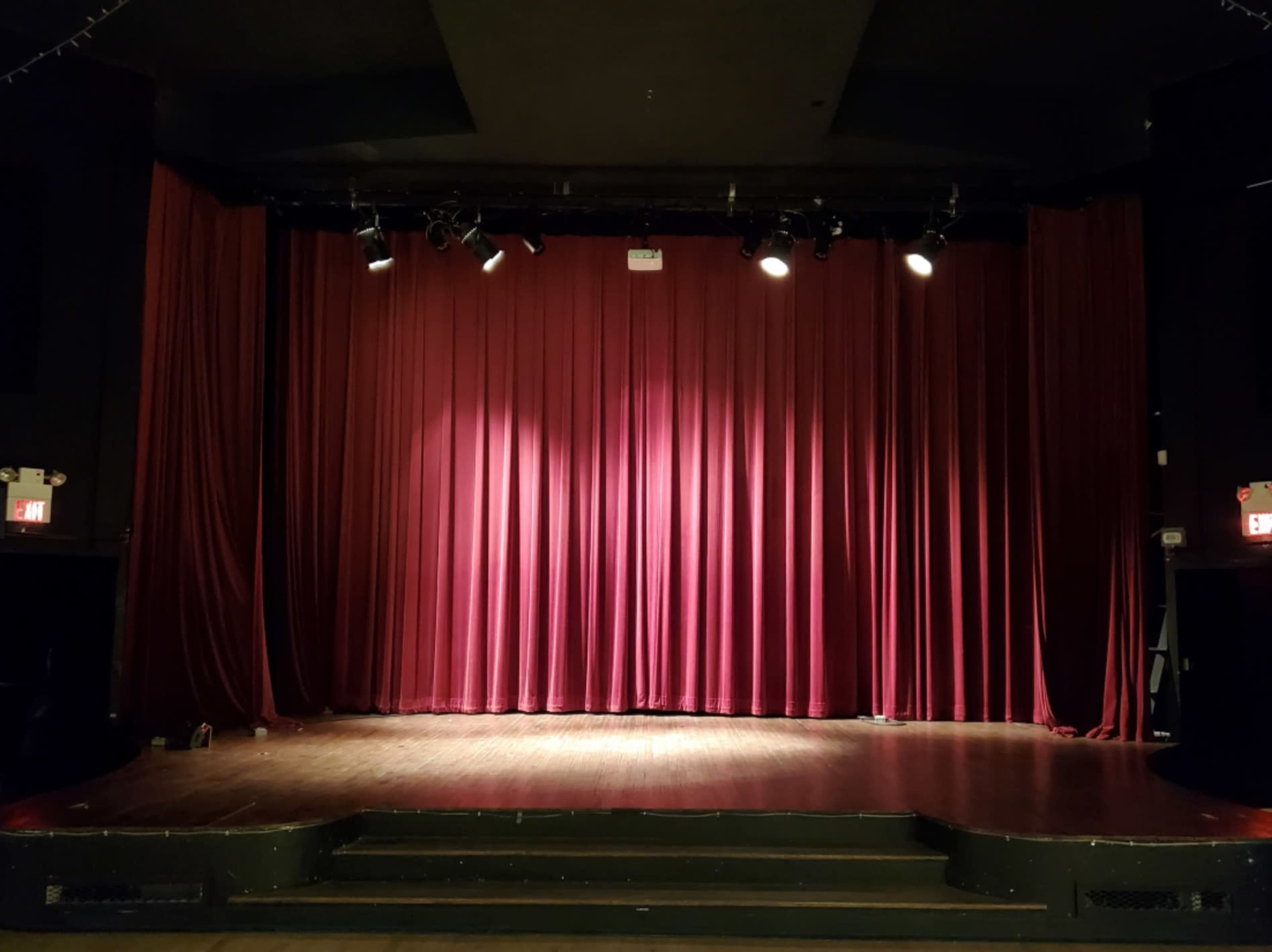The image shows a theater stage with a wooden floor and red curtains, lit by overhead stage lights.