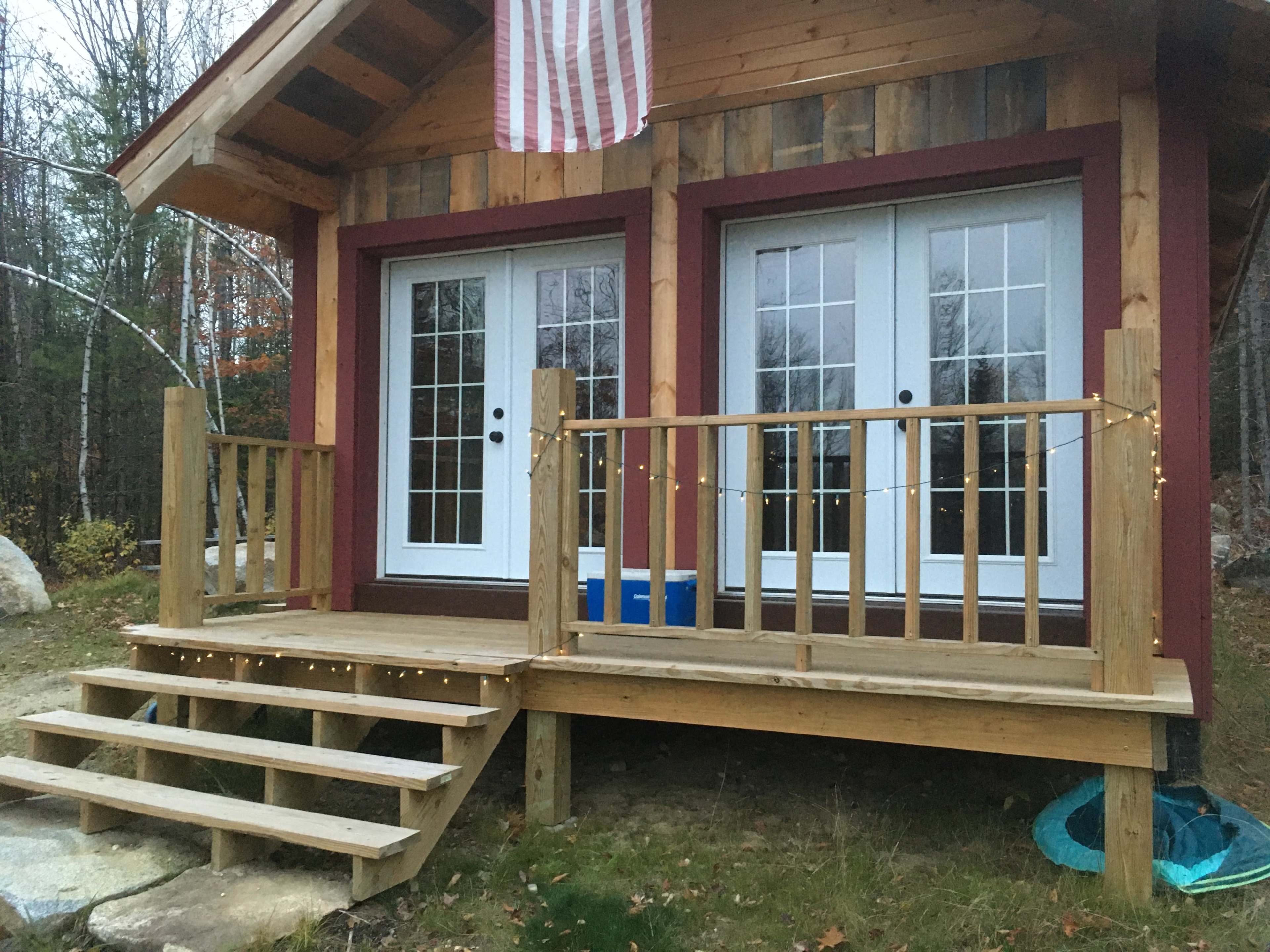 A small wooden cabin features a front porch with a railing, steps leading up to double doors, and an American flag hanging above.