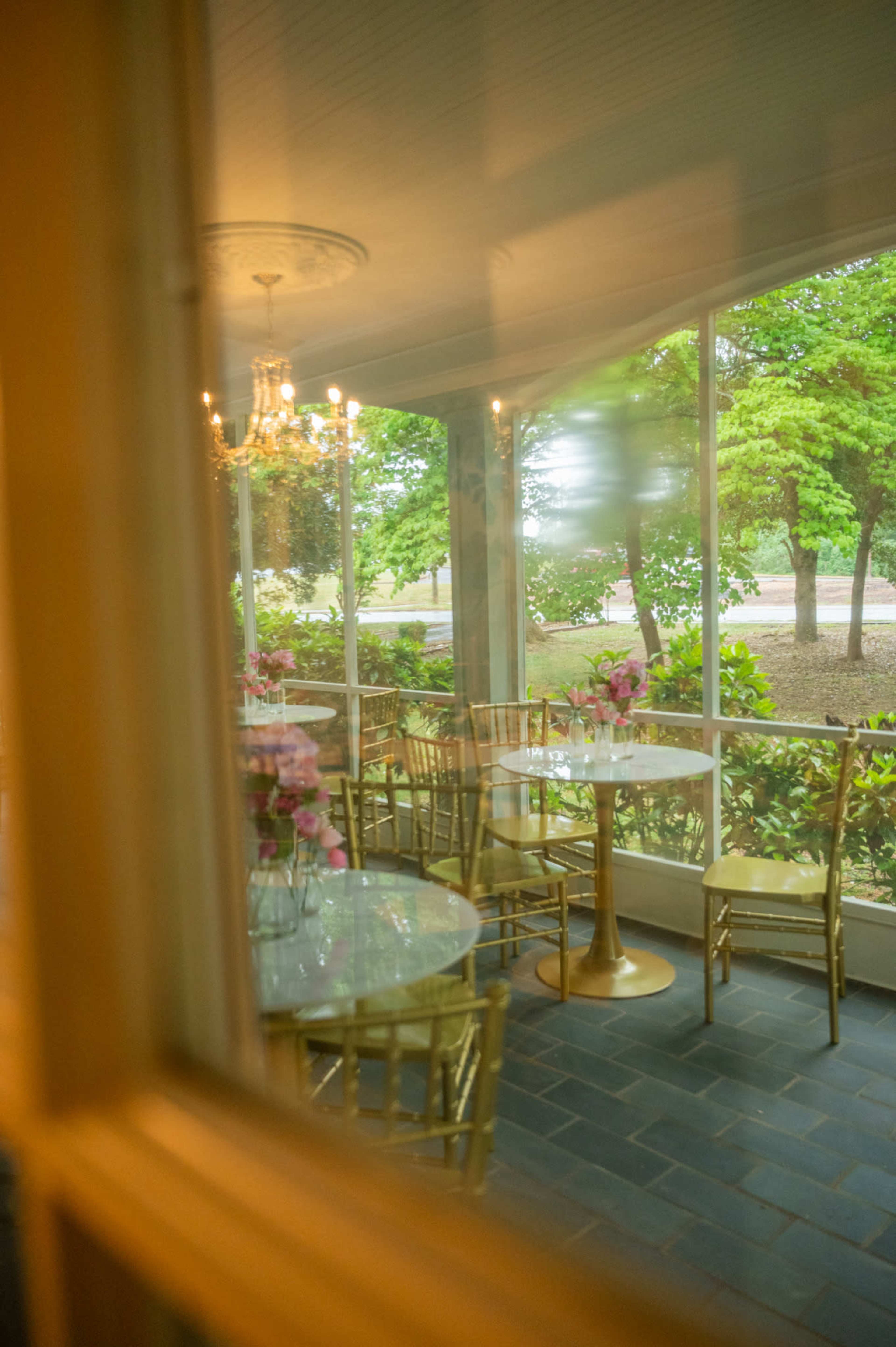 A sunlit enclosed porch features round glass tables and golden chairs, surrounded by greenery.