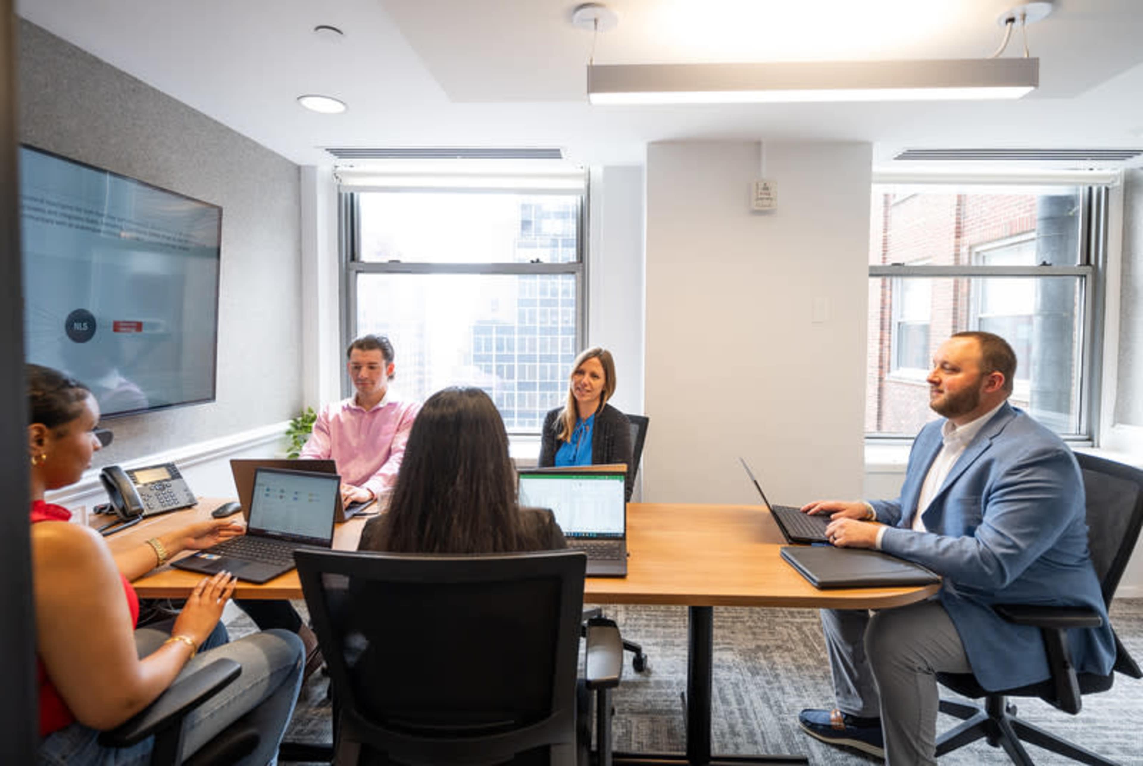 A group of five professionals is seated around a conference table in a modern office meeting room, engaged in discussion while using laptops.