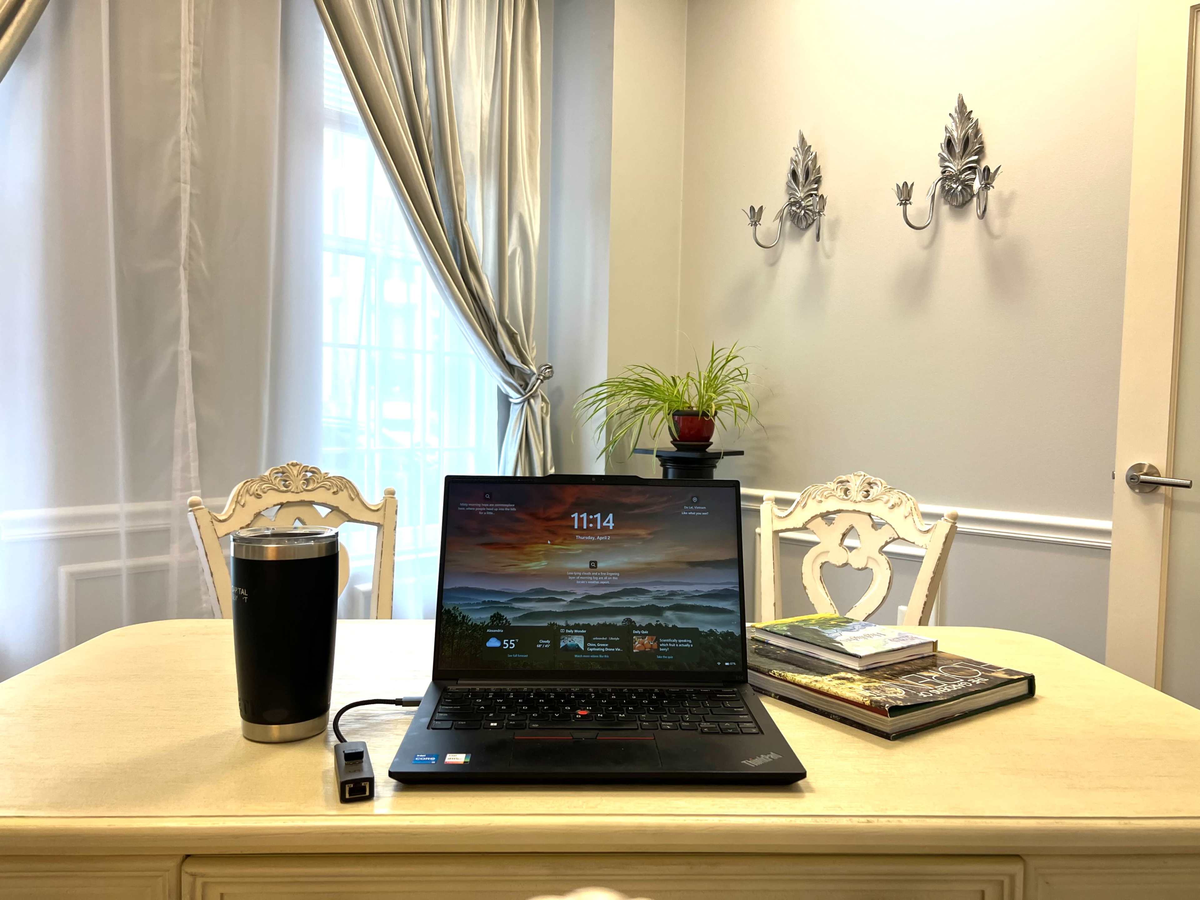 A laptop sits open on a table next to a stainless steel travel mug, with a potted plant and a stack of books in the background.