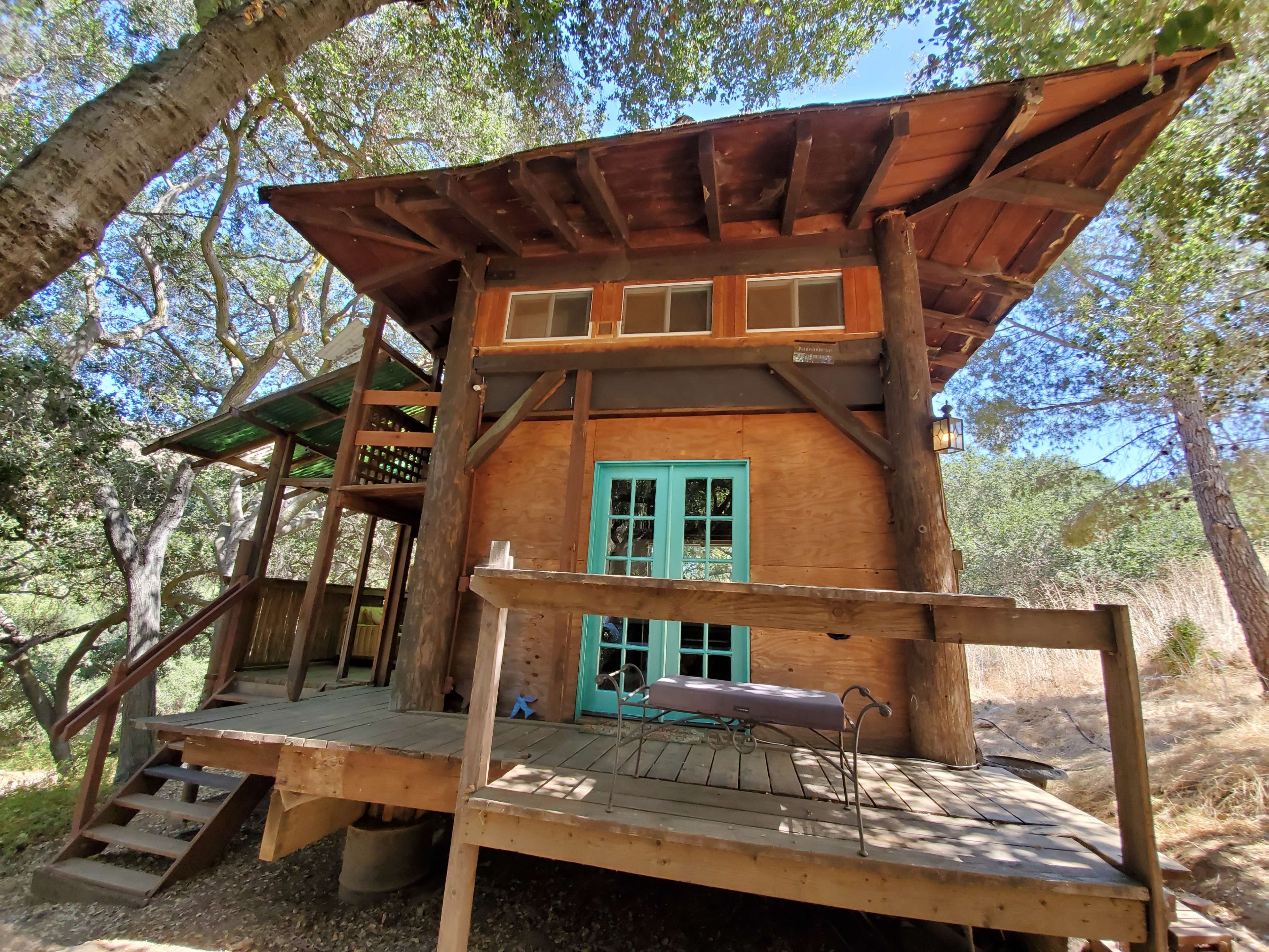 A small wooden cabin with large windows and a covered porch is nestled among trees on a raised platform.