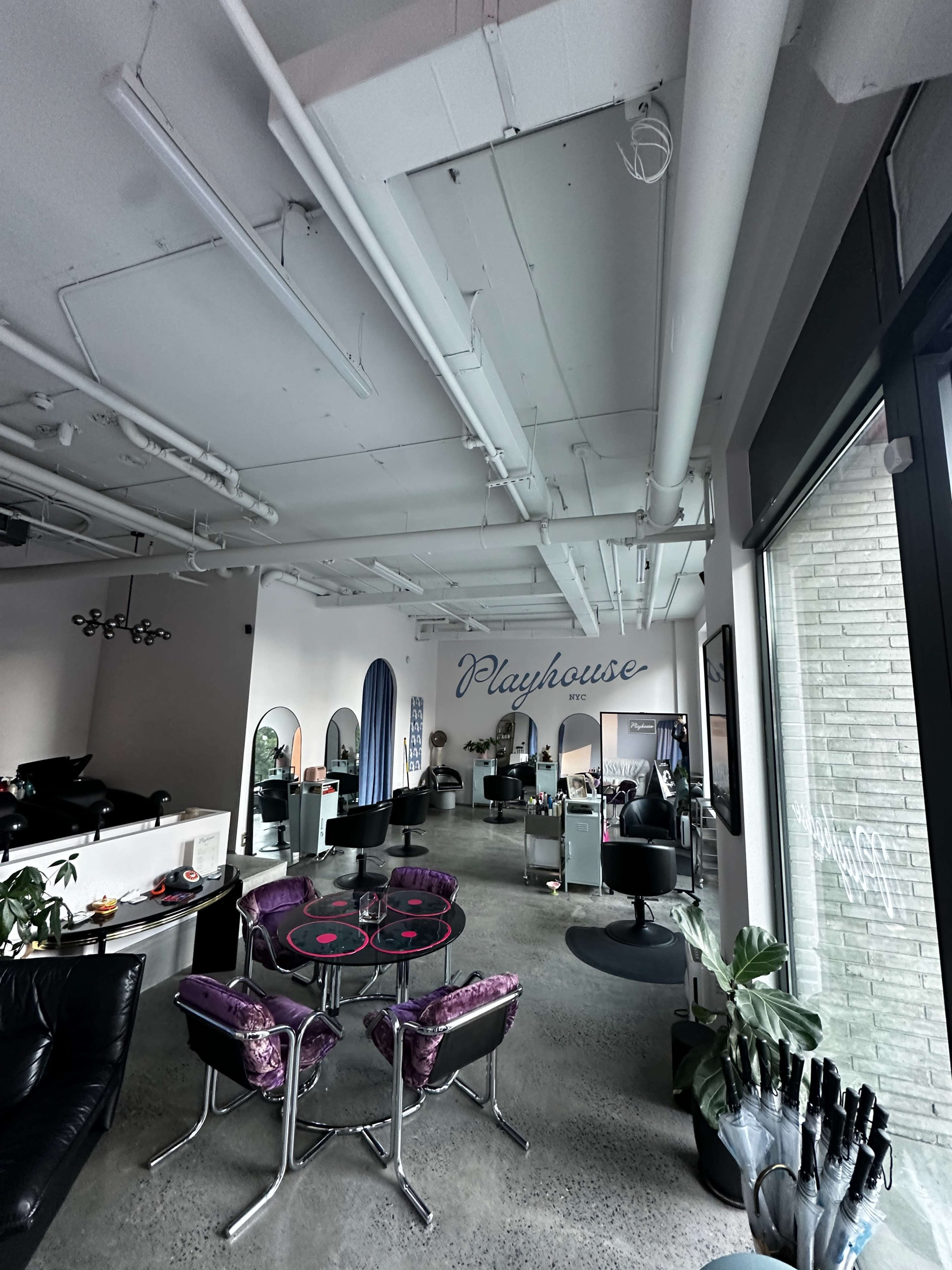 The image shows a spacious salon interior with several styling stations, a circular table with chairs, and a sign that reads "Playhouse" on the wall.