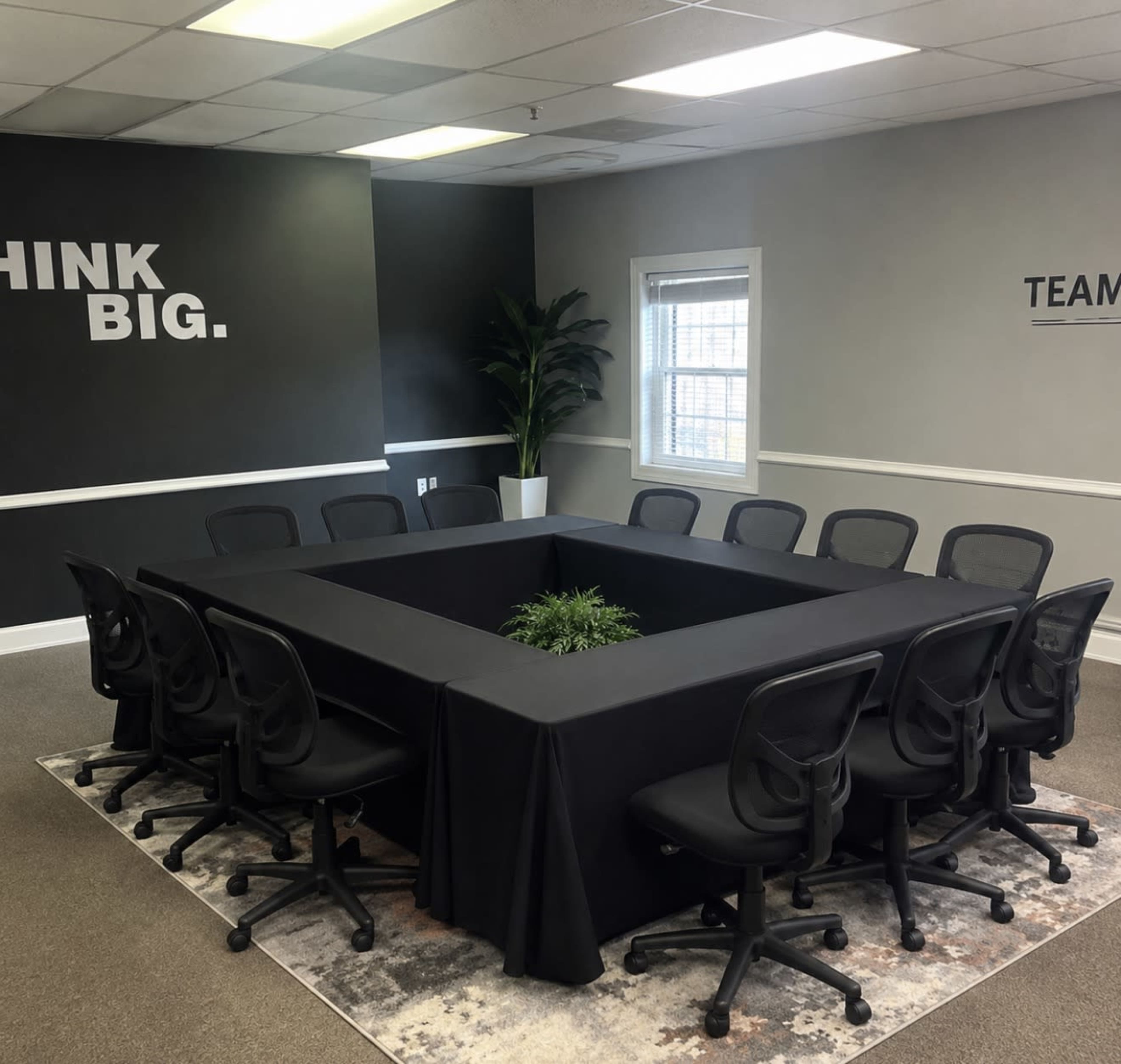 The image shows a conference room with a large black table surrounded by twelve ergonomic chairs, featuring a small plant in the center and motivational text on the walls.