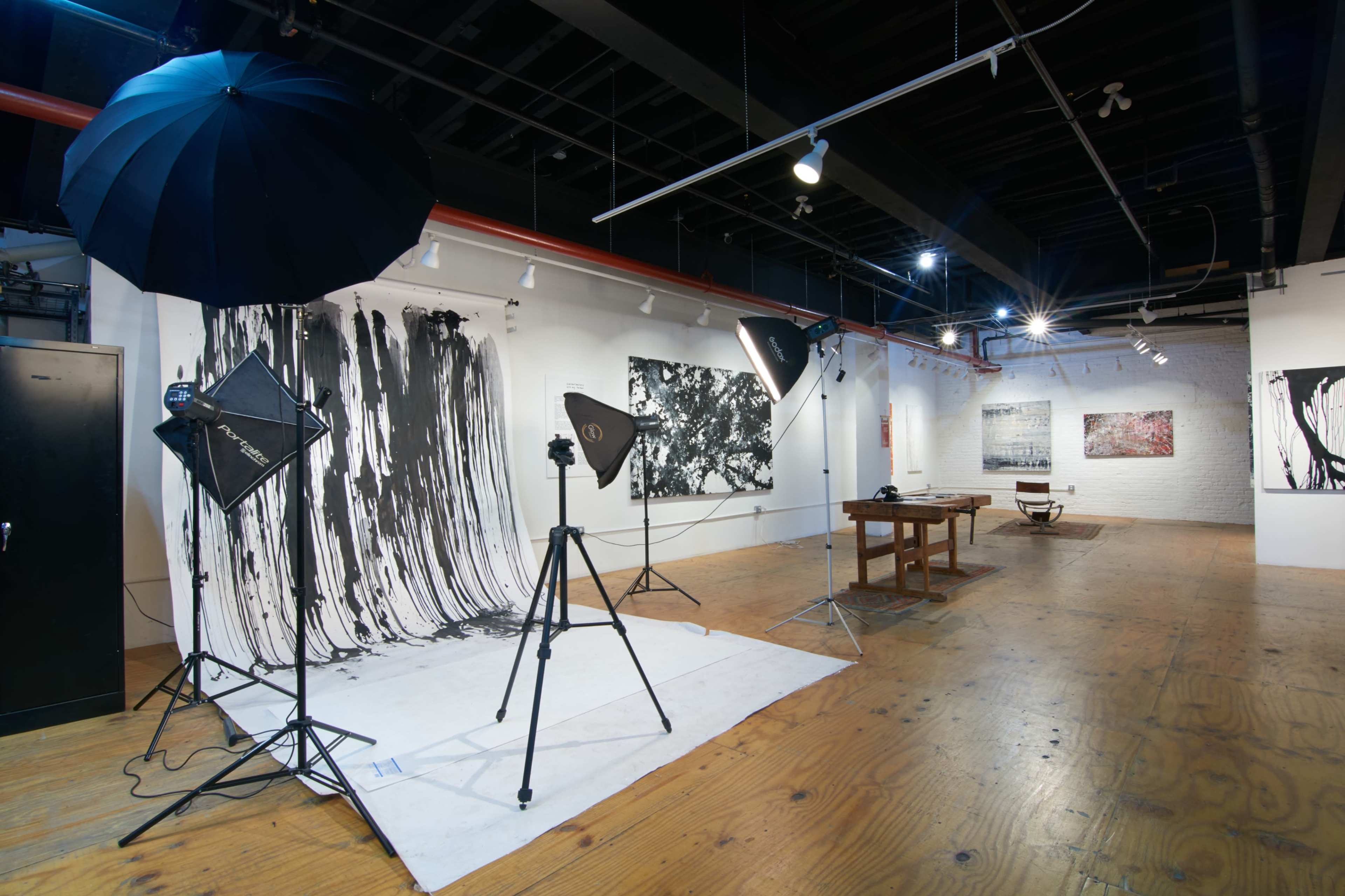 The image shows an art gallery featuring large black-and-white abstract paintings, photography equipment, and a wooden table with a chair in the center.