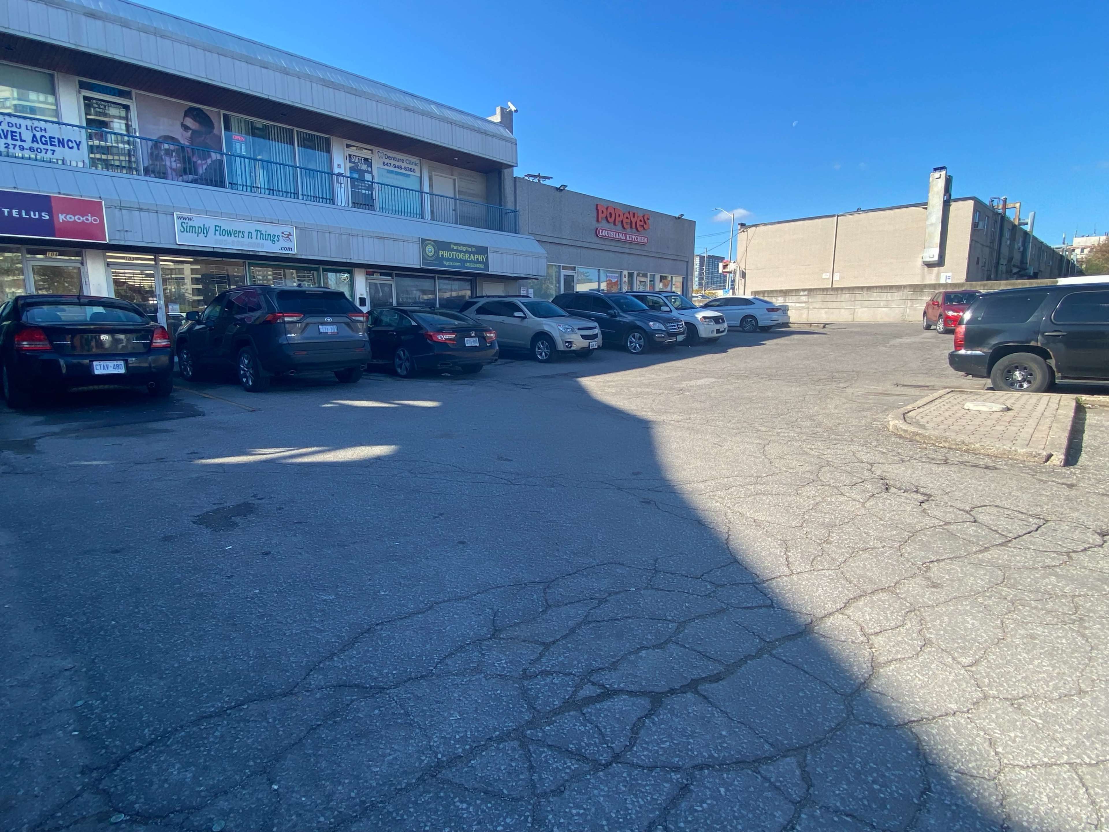 A parking lot next to a row of businesses, with various parked cars and clear blue skies overhead.