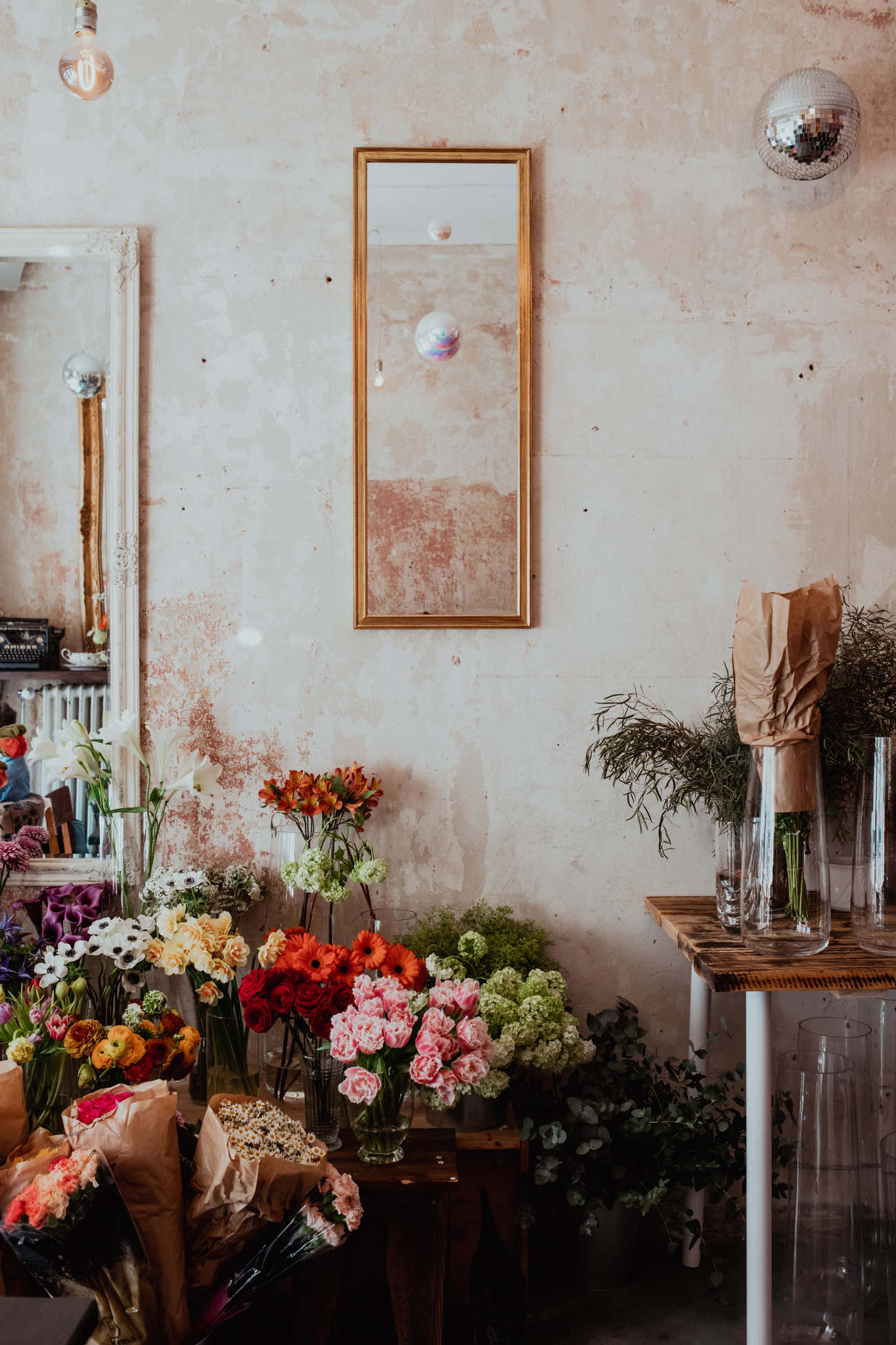 The image shows a flower shop with various colorful arrangements of flowers displayed near a textured wall, along with a mirror reflecting light and decor.