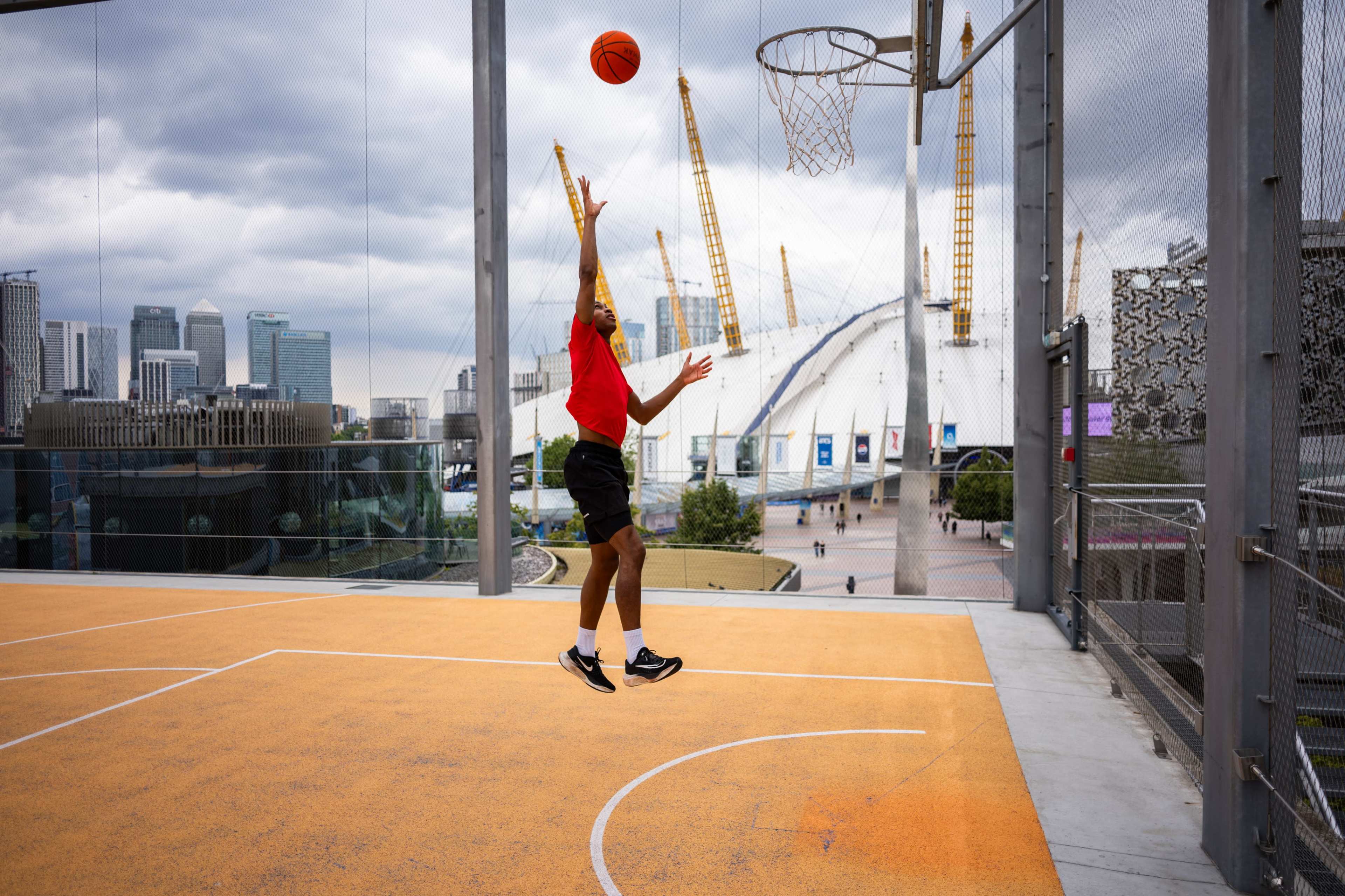 A player jumps to shoot a basketball towards a hoop on a rooftop court with a city skyline in the background.