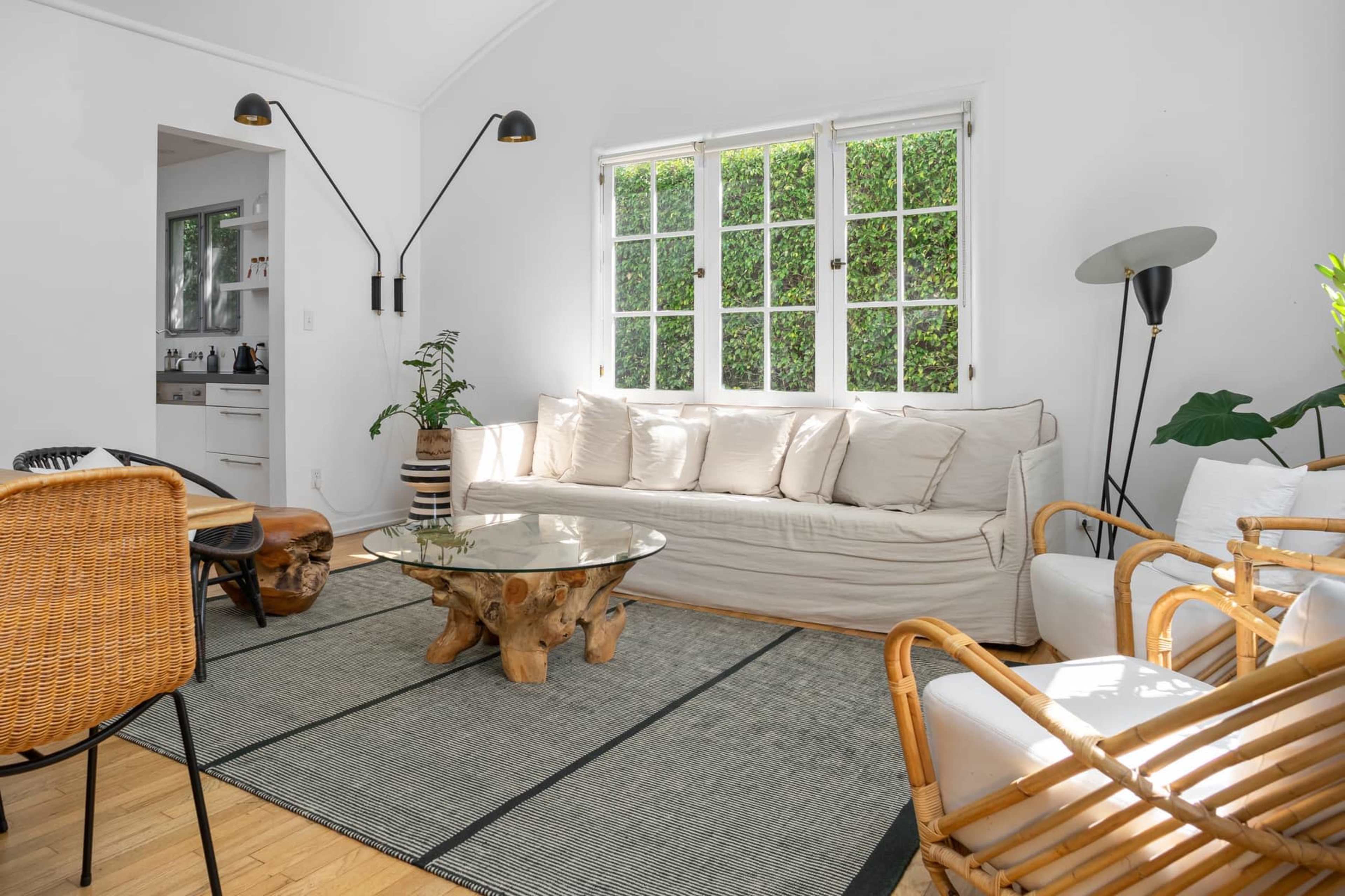 The living room features a light-colored sofa, a round glass coffee table made from wood, and various seating arrangements around a natural fiber rug, with large windows allowing sunlight to enter.