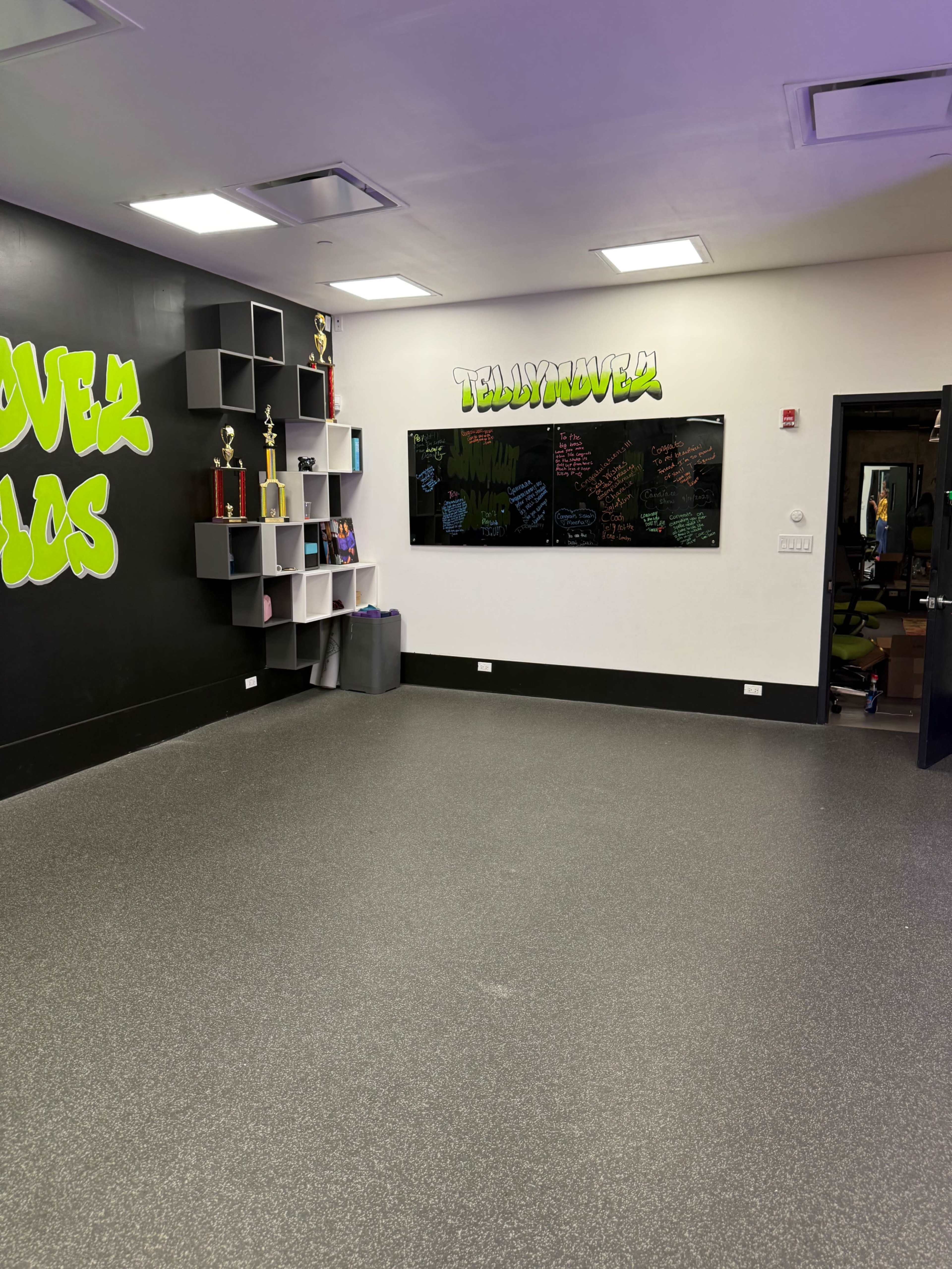 The image shows an empty room with black walls featuring green graffiti-style lettering, shelves with trophies, and a whiteboard covered in colorful writing.