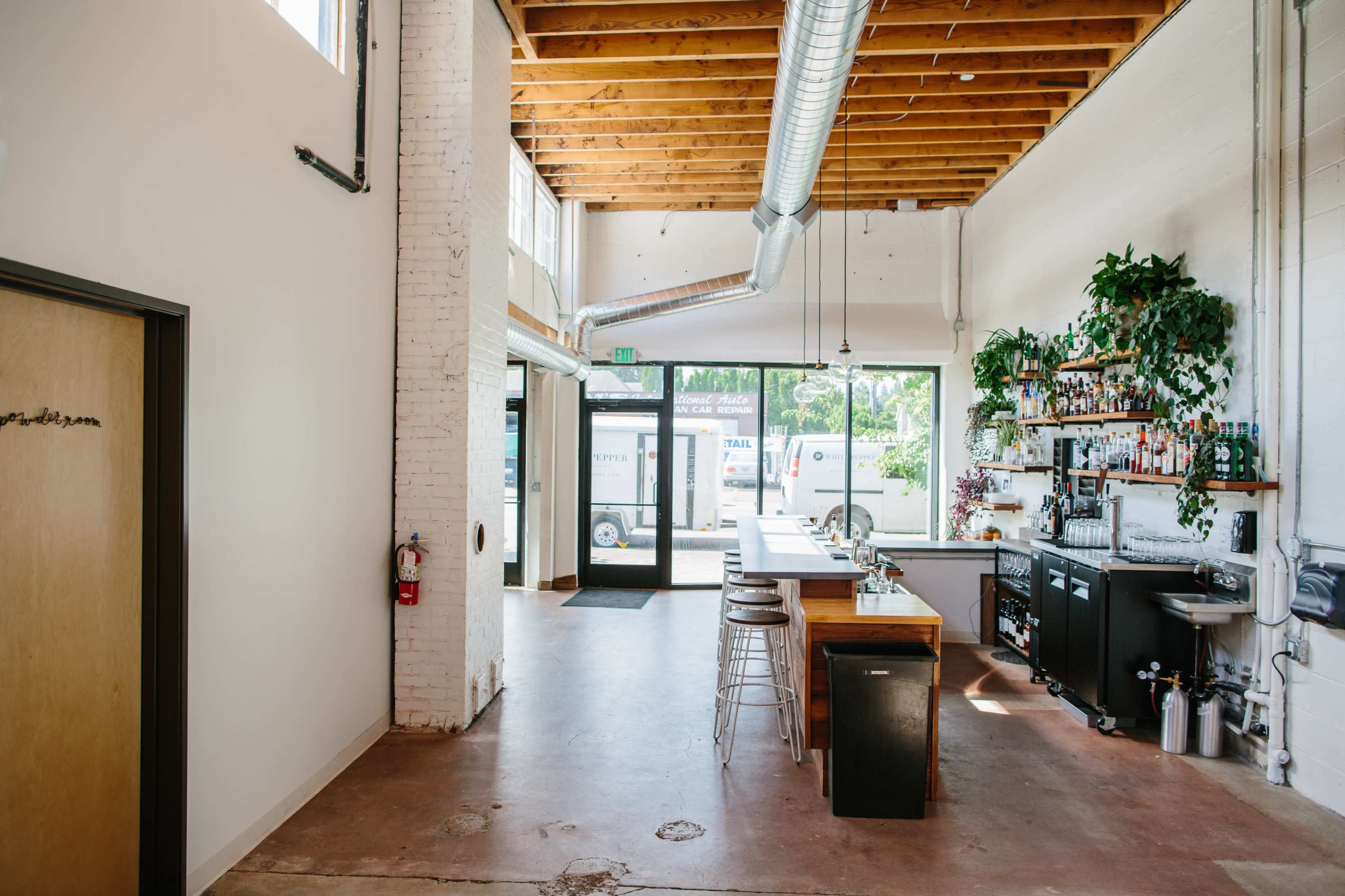A modern open space with a high ceiling, featuring a bar area on the left, large windows, and plants along the walls.