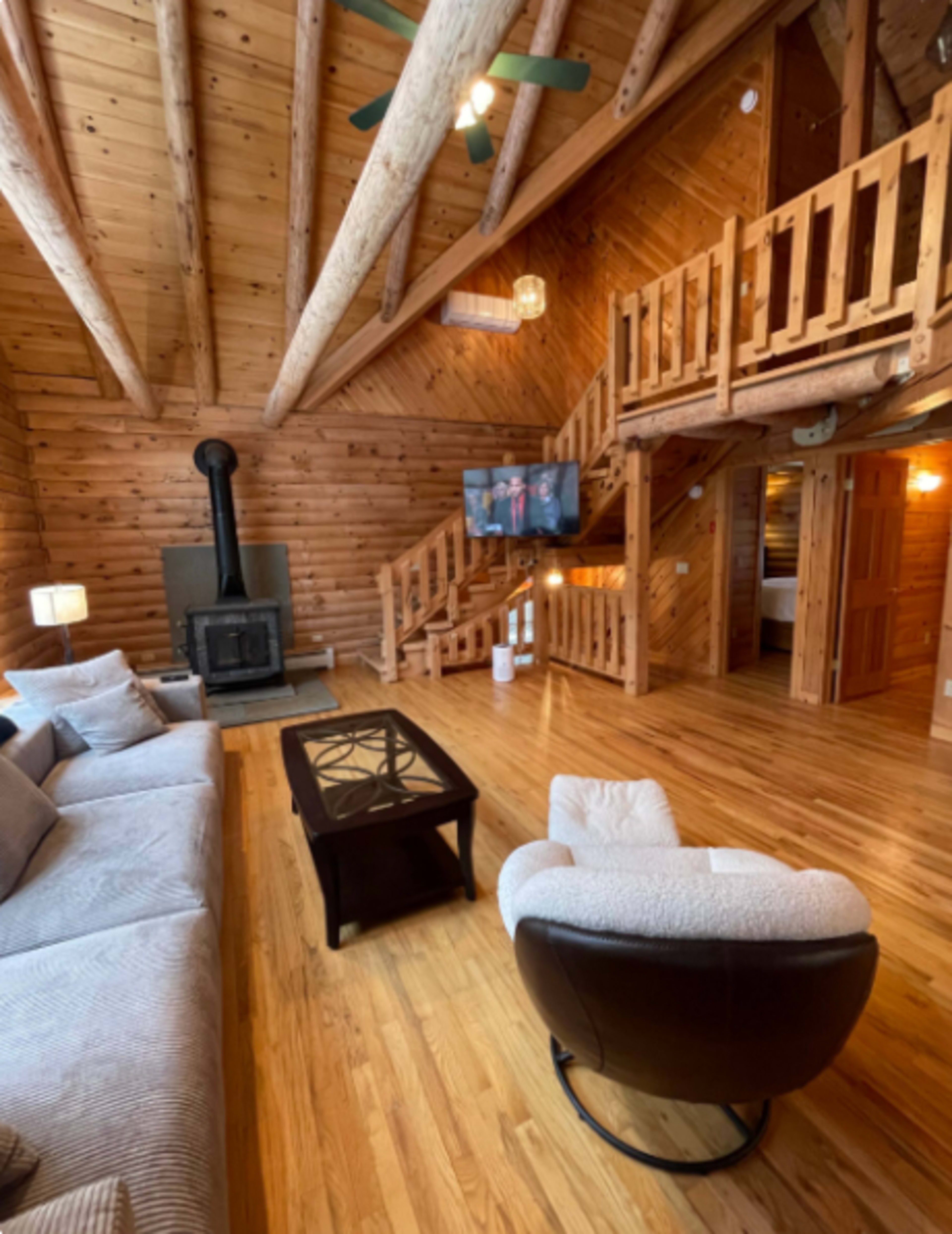 The image shows a cozy wooden cabin living room with a fireplace, a sectional sofa, and an upper loft area connected by a staircase.