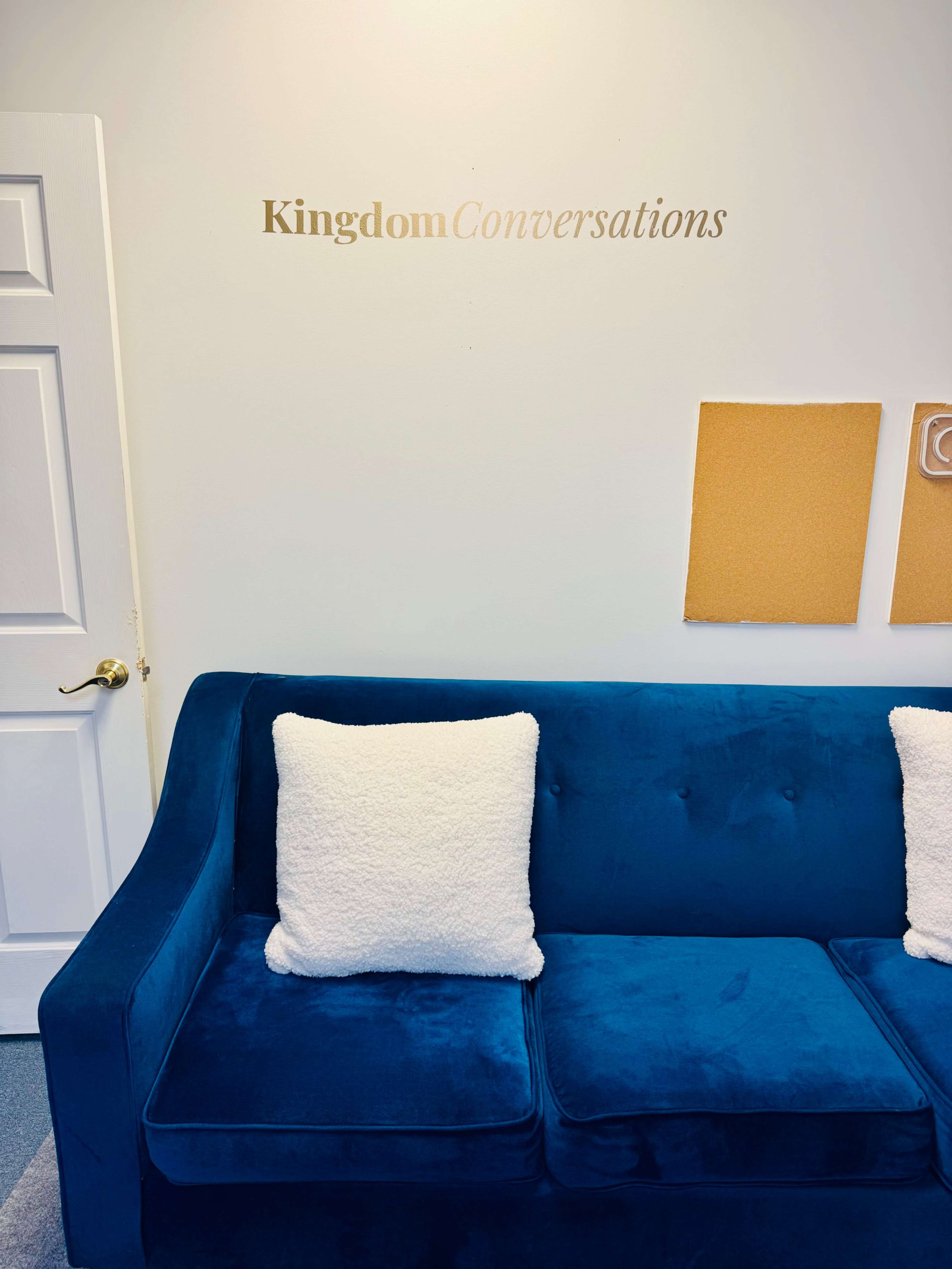 A blue couch with two white pillows sits against a wall displaying the words "Kingdom Conversations" and two cork boards nearby.