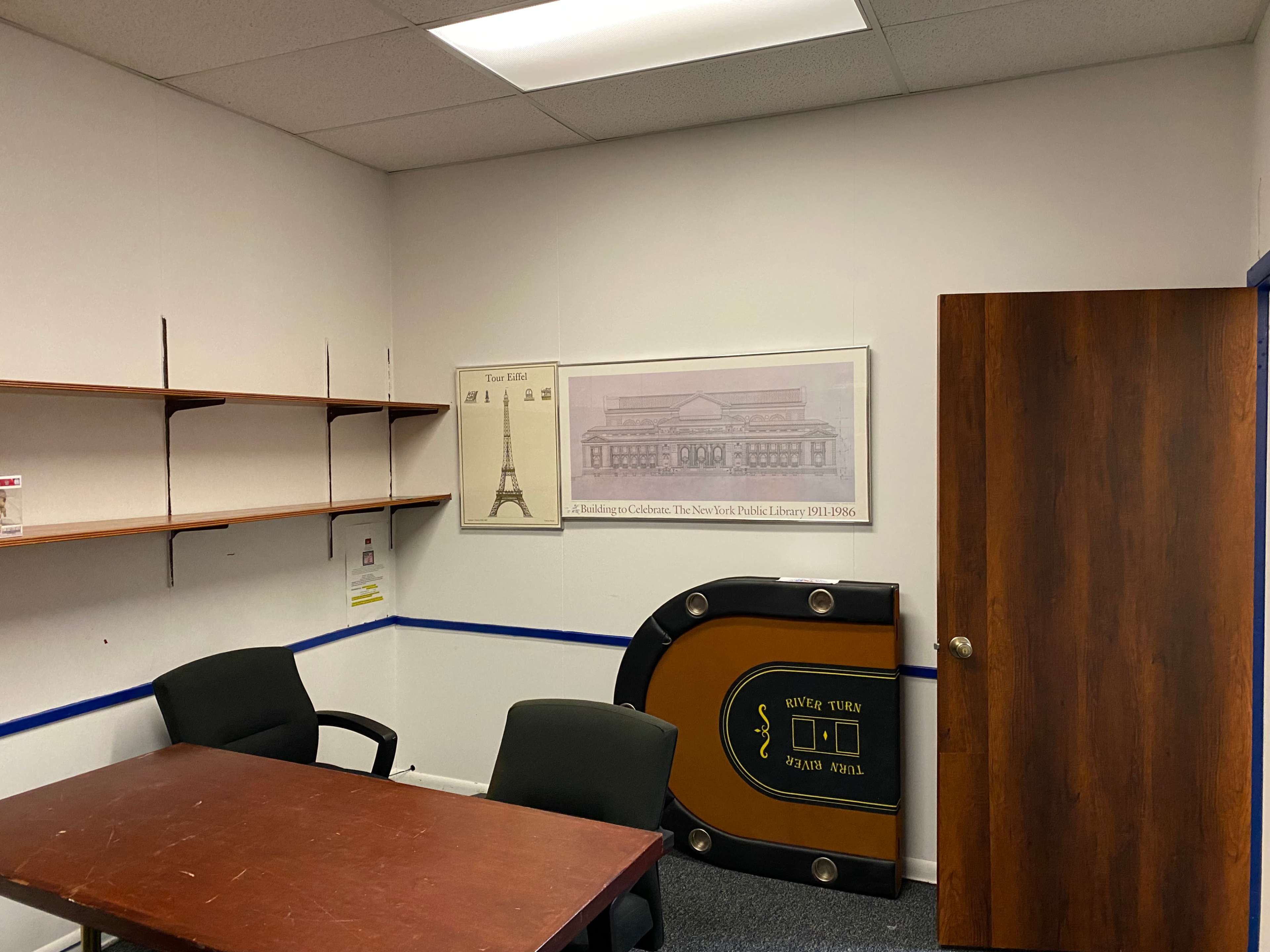 The image shows a small office with a wooden table, two black chairs, a framed poster on the wall, a shelf, and a door.