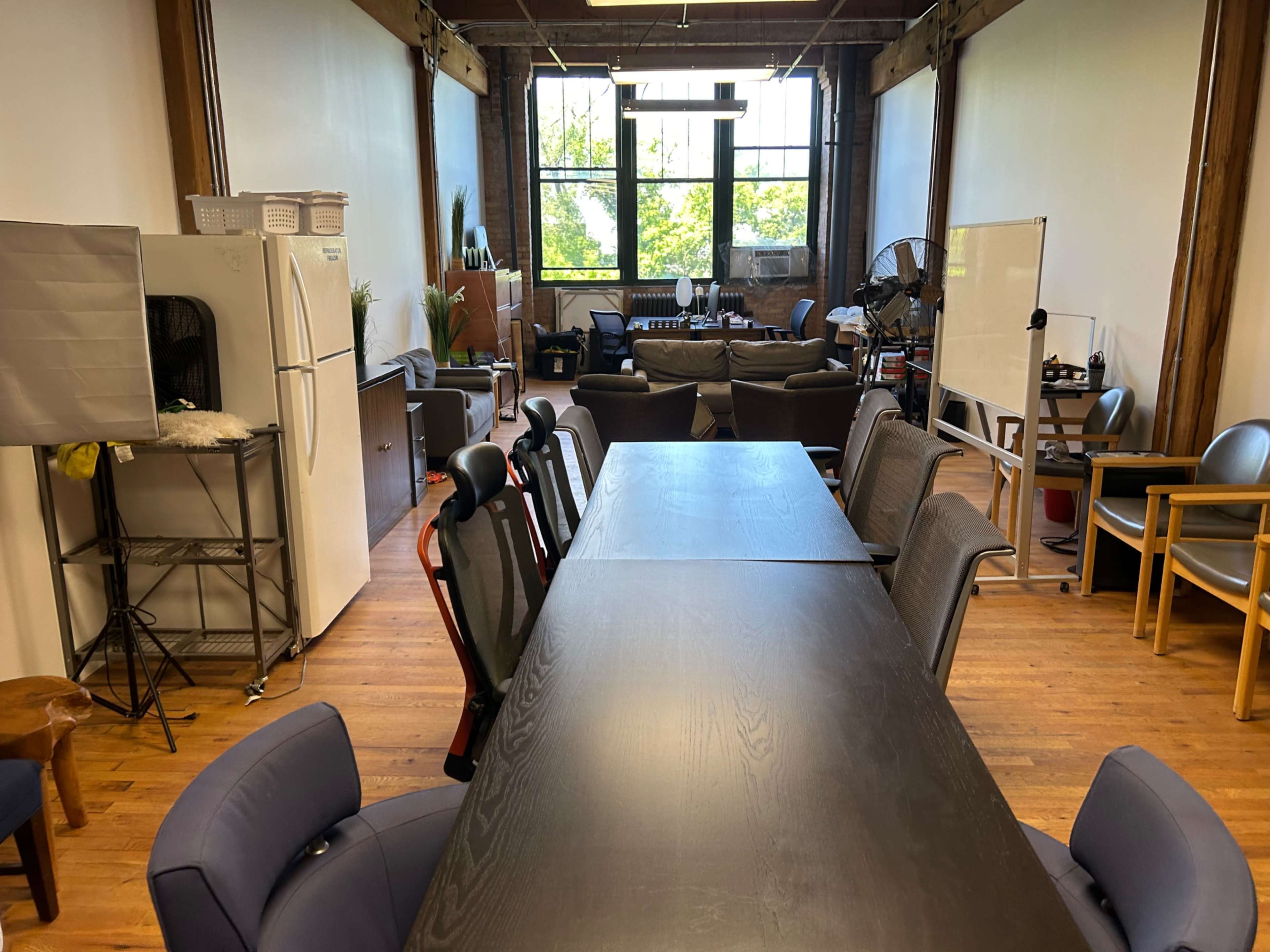 The image shows a spacious office setting with a long black table surrounded by chairs, a seating area with sofas in the background, and large windows letting in natural light.