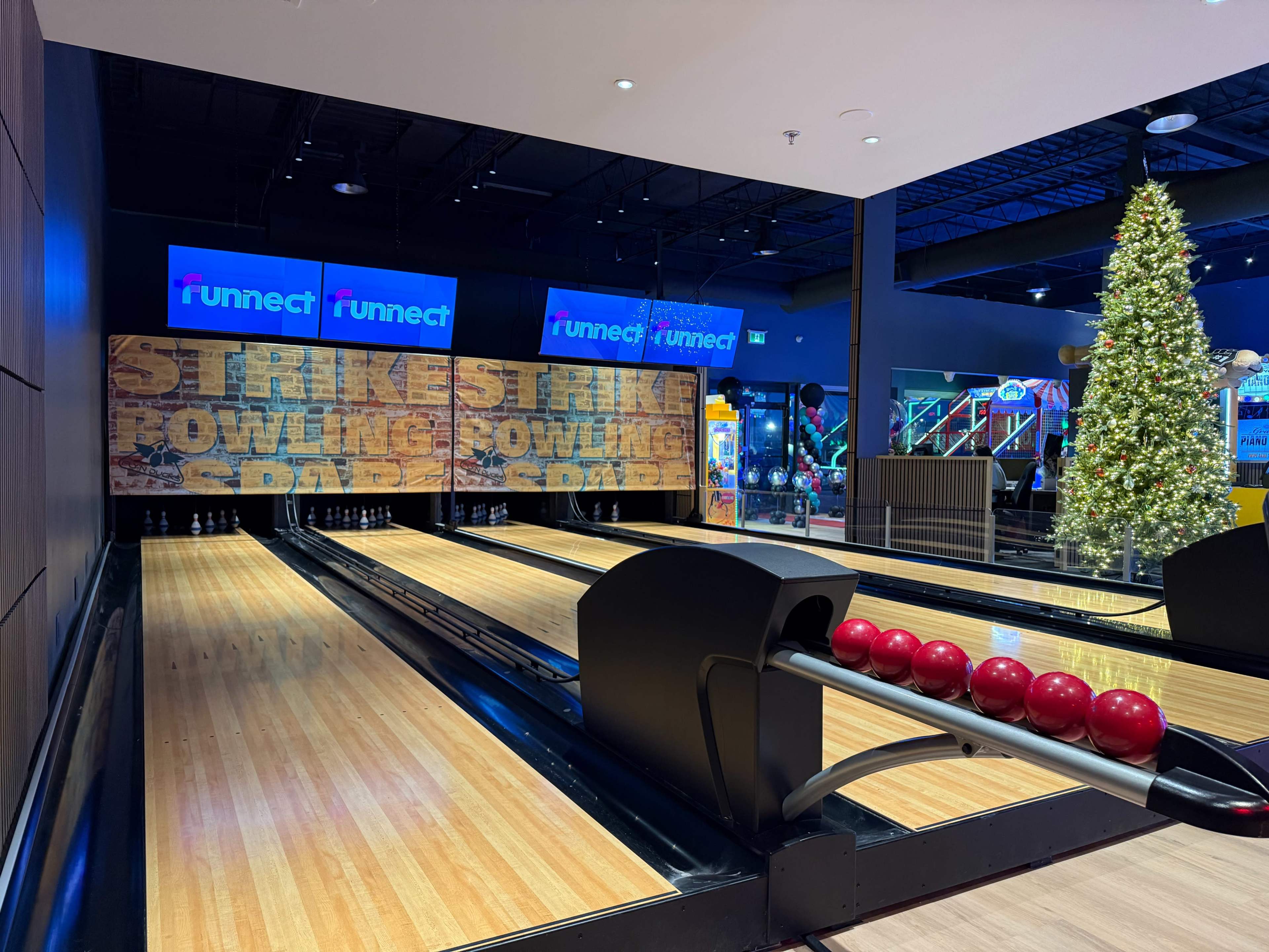 The image shows a bowling area with two lanes, bright red bowling balls, and a large decorative Christmas tree in the background.