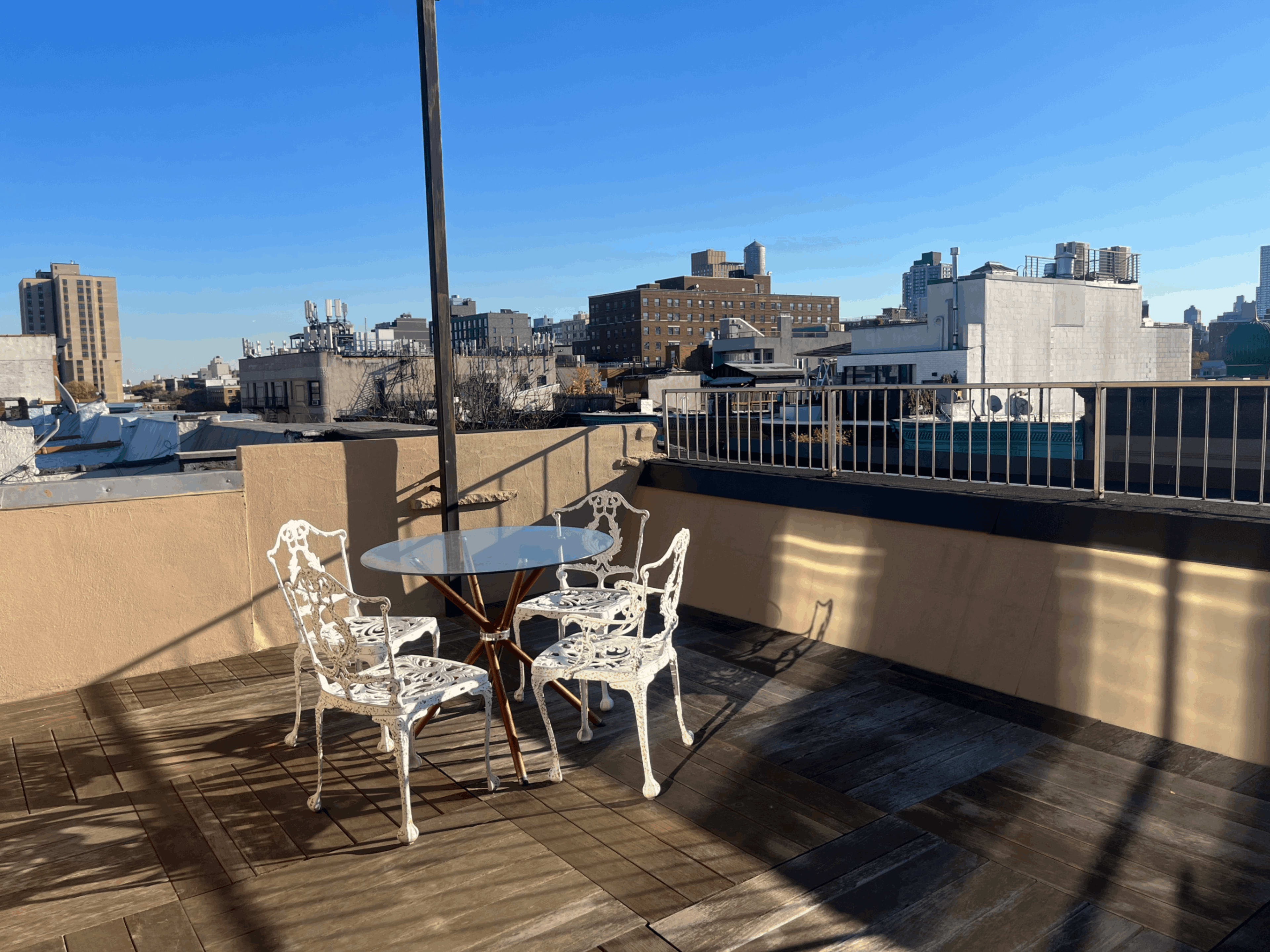 A rooftop patio with a glass table and four white metal chairs set against a backdrop of urban buildings and a clear blue sky.