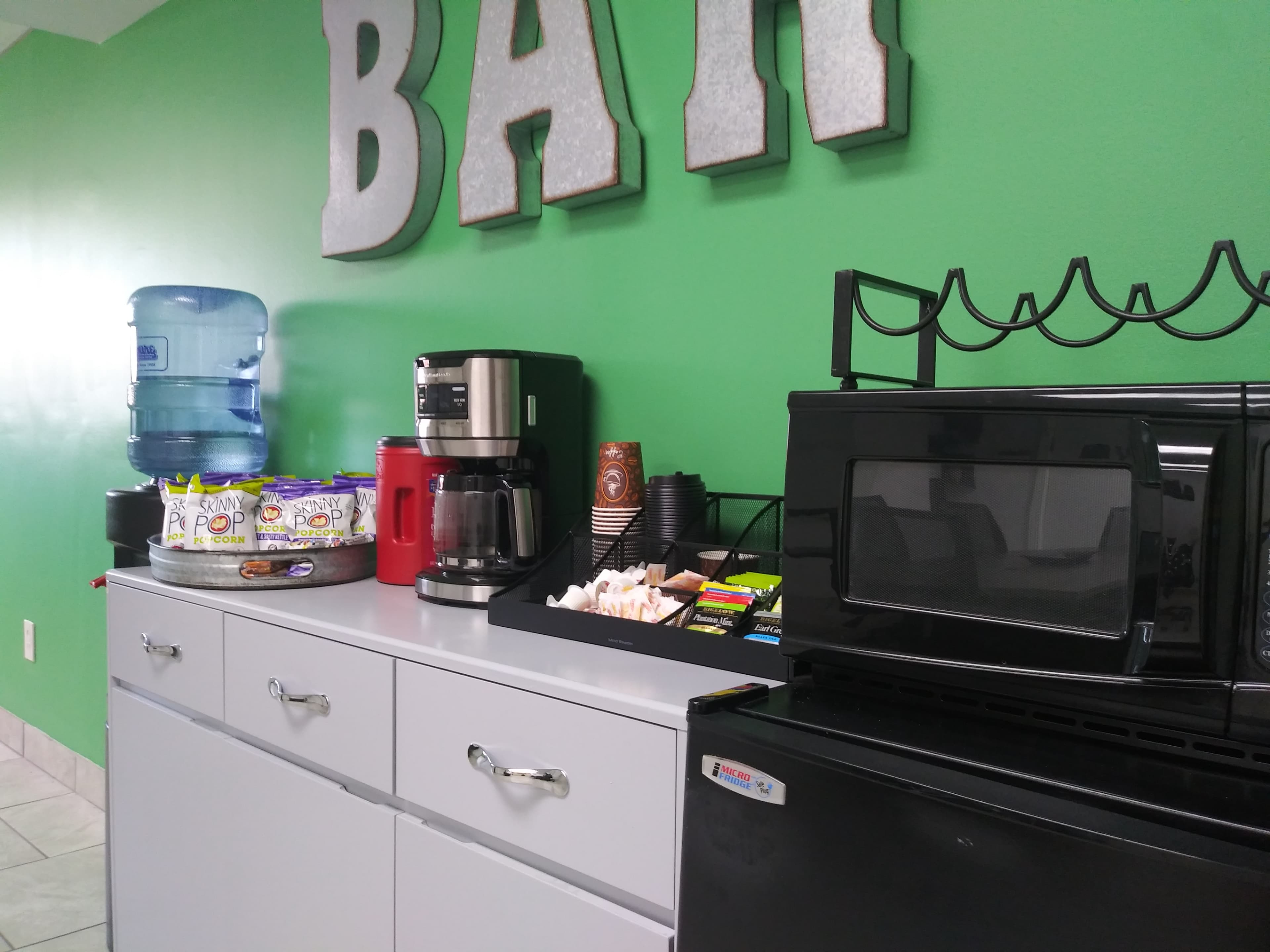 A kitchenette area features a water cooler, coffee maker, microwave, and assorted snacks on a countertop in front of a green wall with a sign that says "BAR."