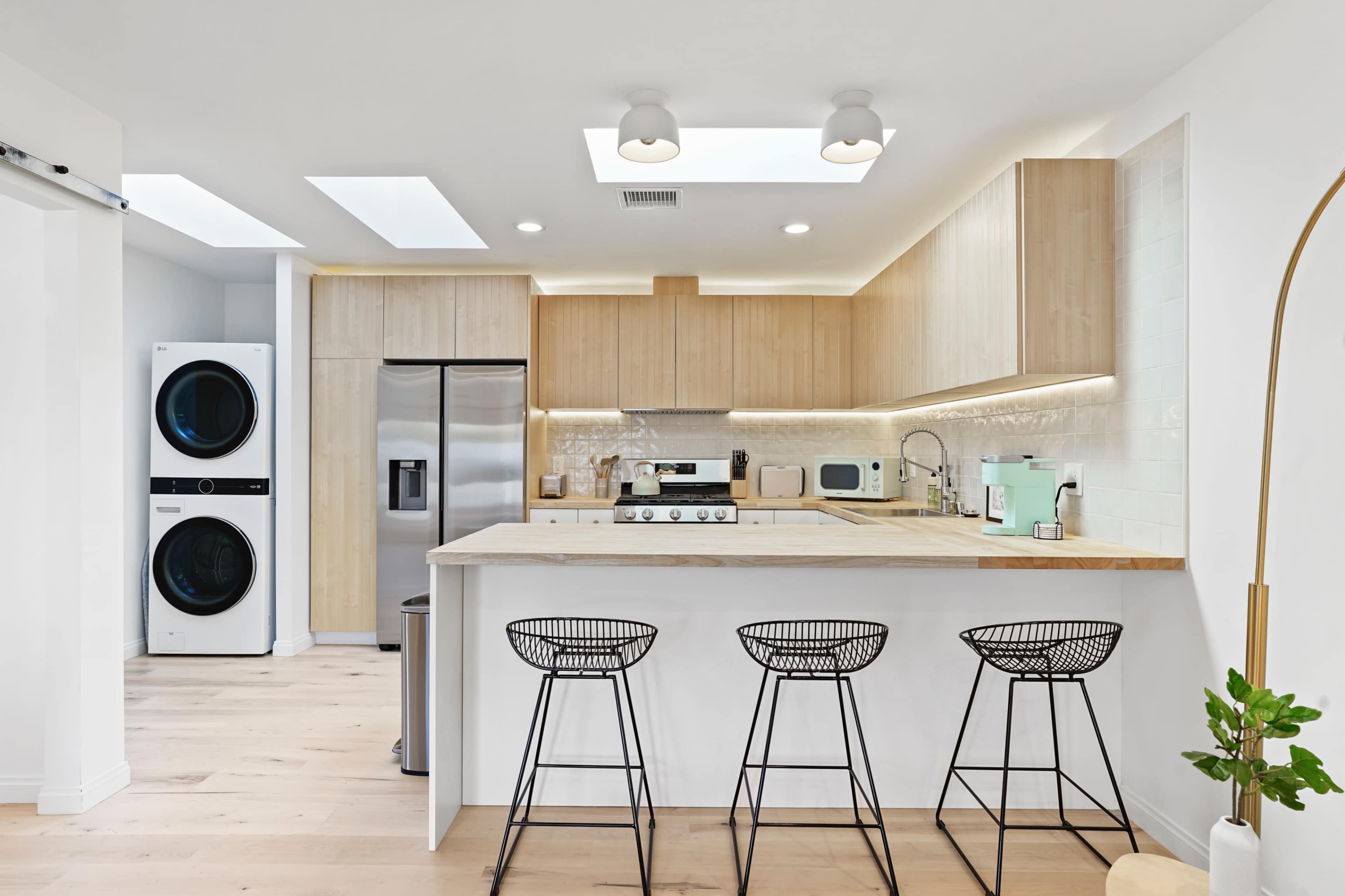 A modern kitchen features wooden cabinetry, stainless steel appliances, and a bar with three wire stools, alongside a laundry area with stacked washer and dryer units.