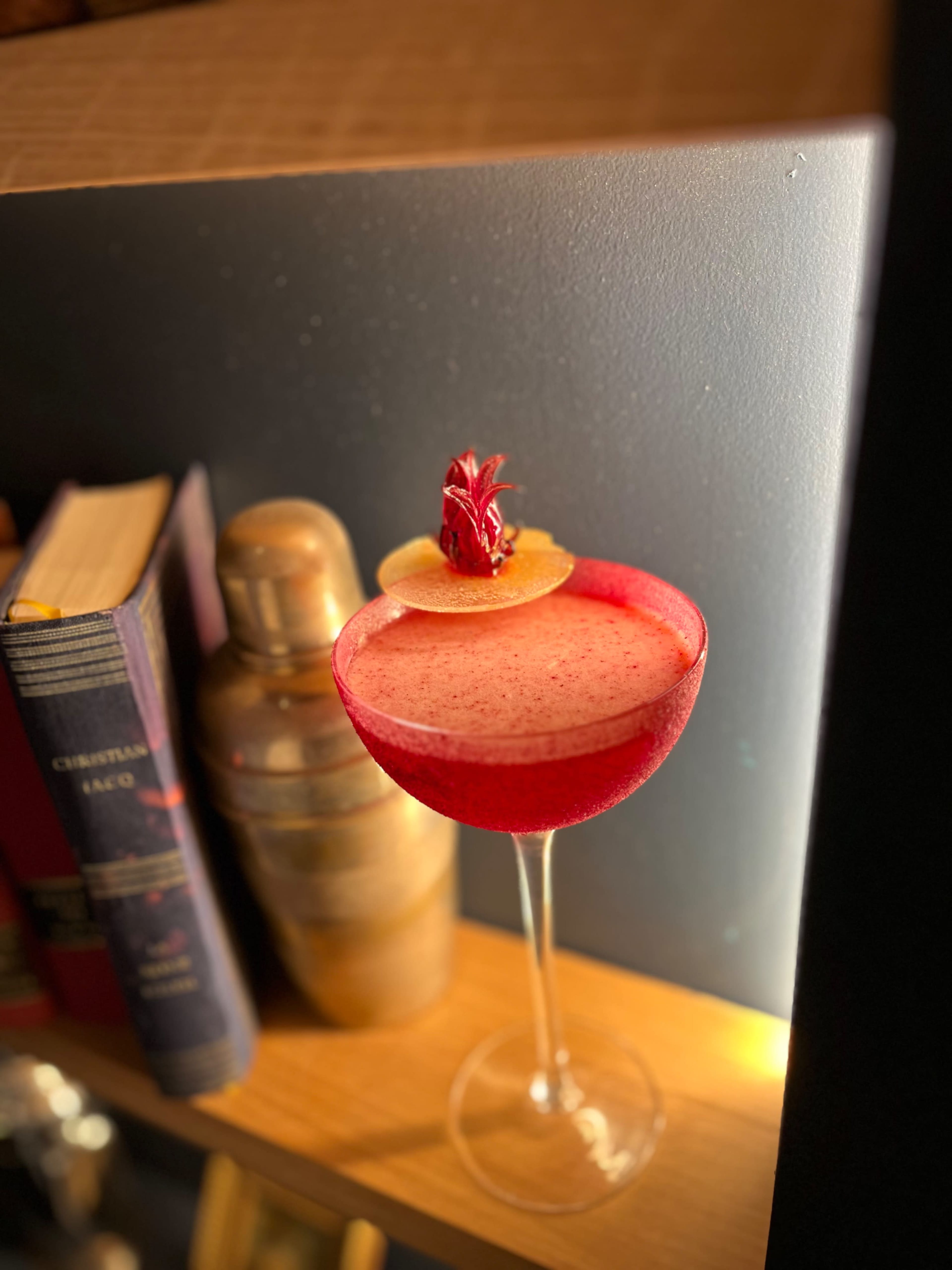 A vibrant cocktail is placed on a wooden shelf next to a shaker and books.