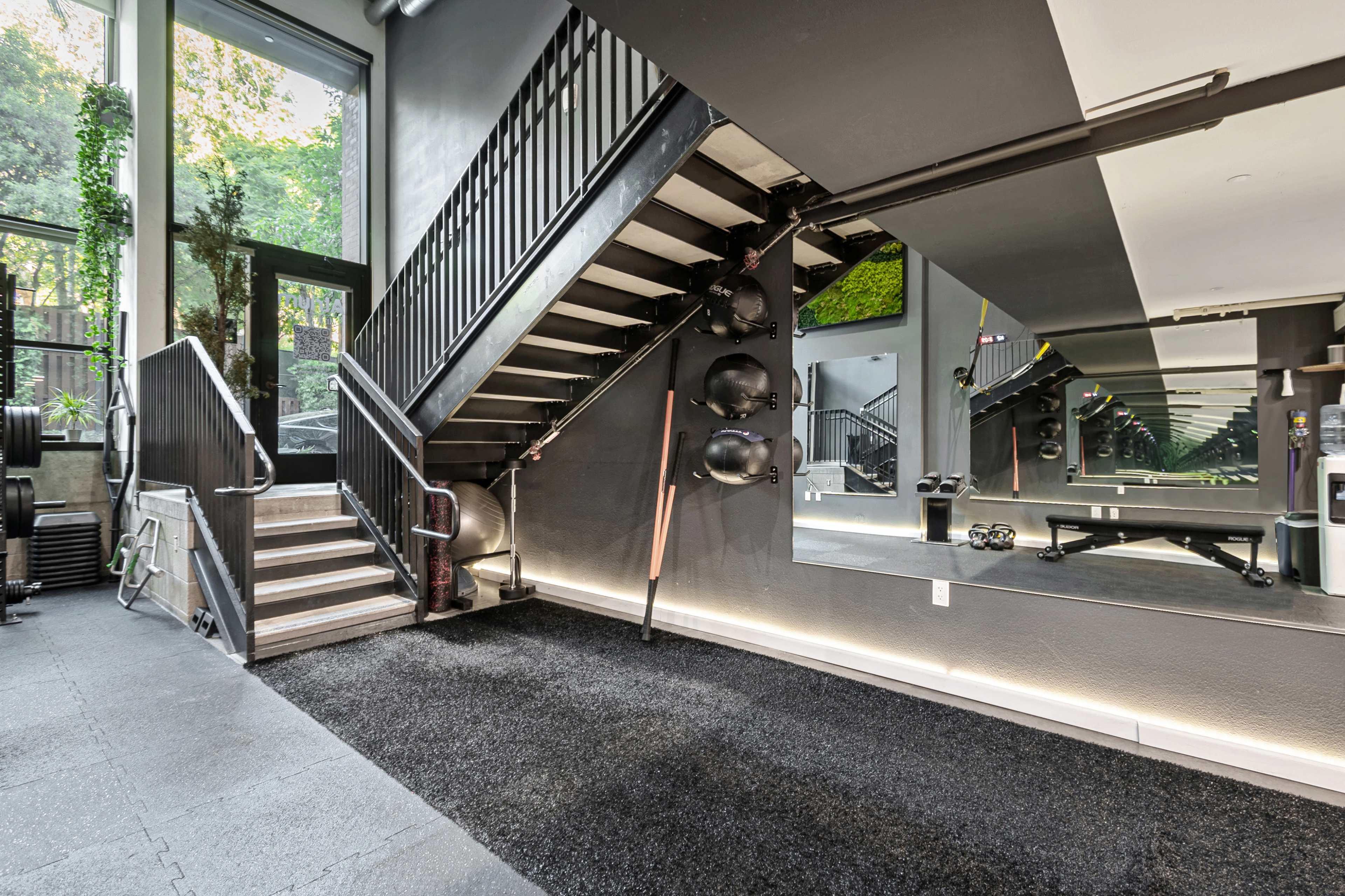 A modern gym space with a staircase, workout equipment, and a mirrored wall, located beneath a large window with greenery visible outside.