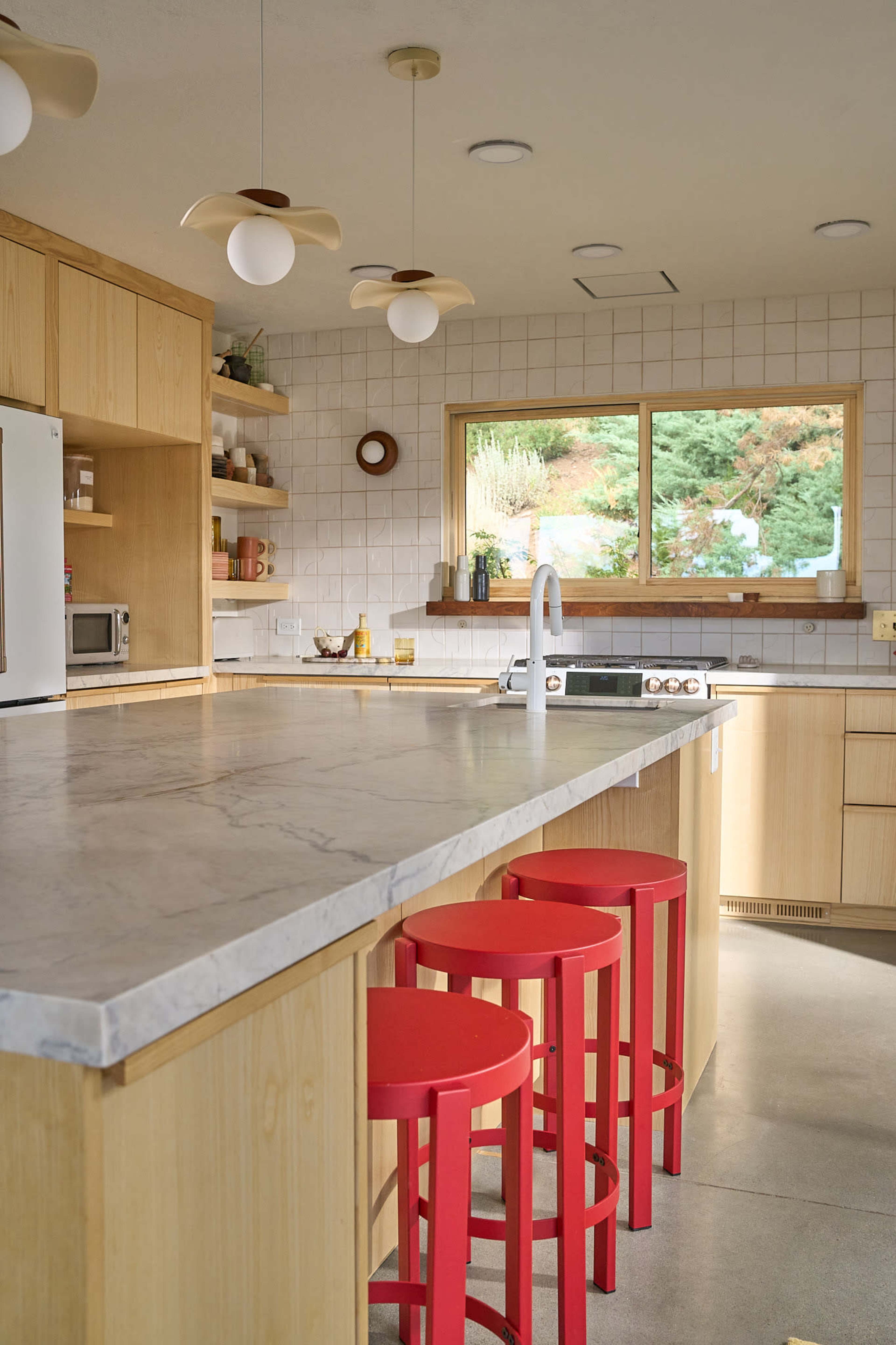 The image shows a modern kitchen featuring a marble countertop island, wooden cabinetry, and three red stools.