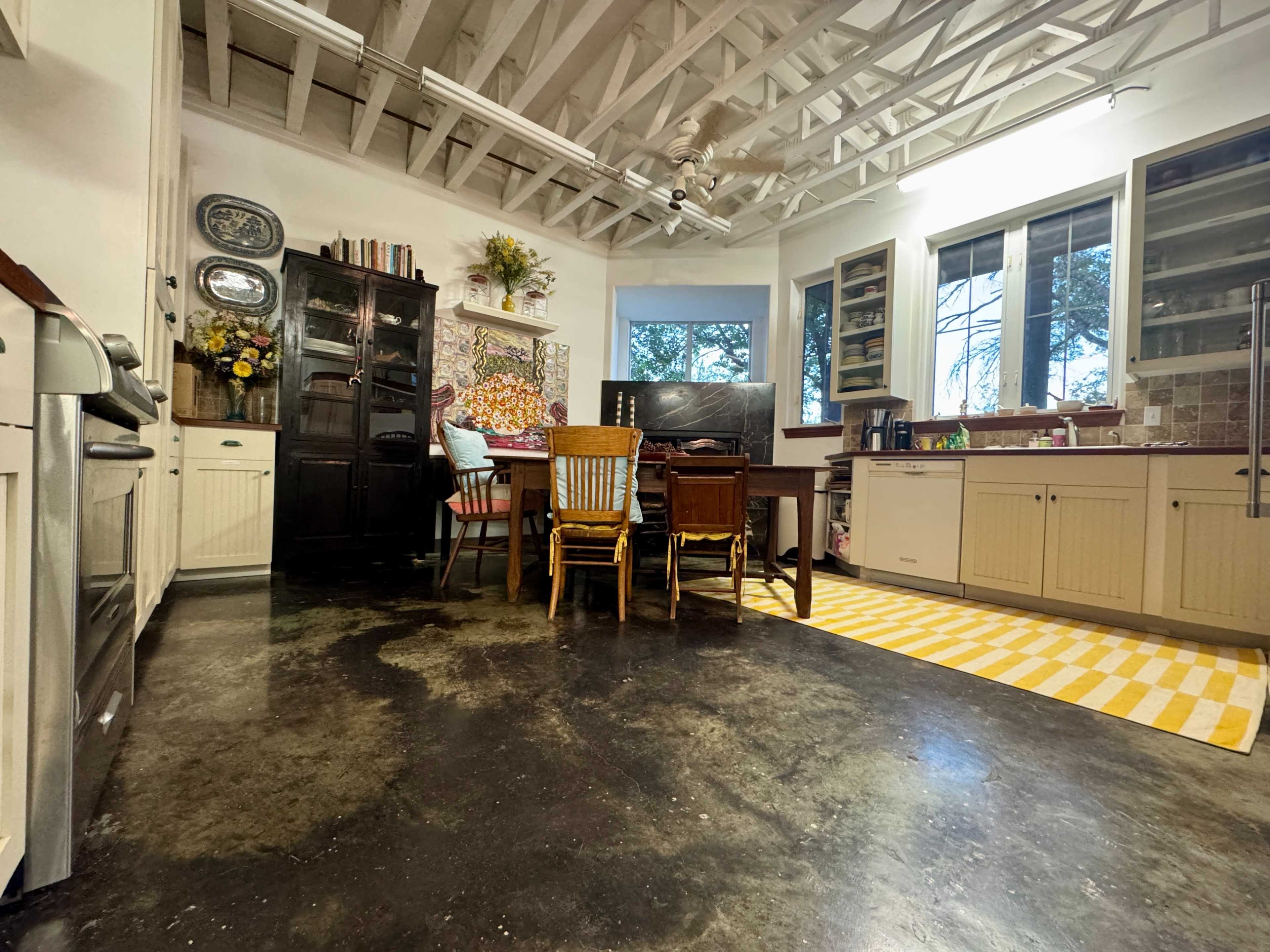 The image shows a spacious kitchen with a wooden dining table surrounded by chairs, a large window letting in natural light, and a yellow striped rug on the floor.