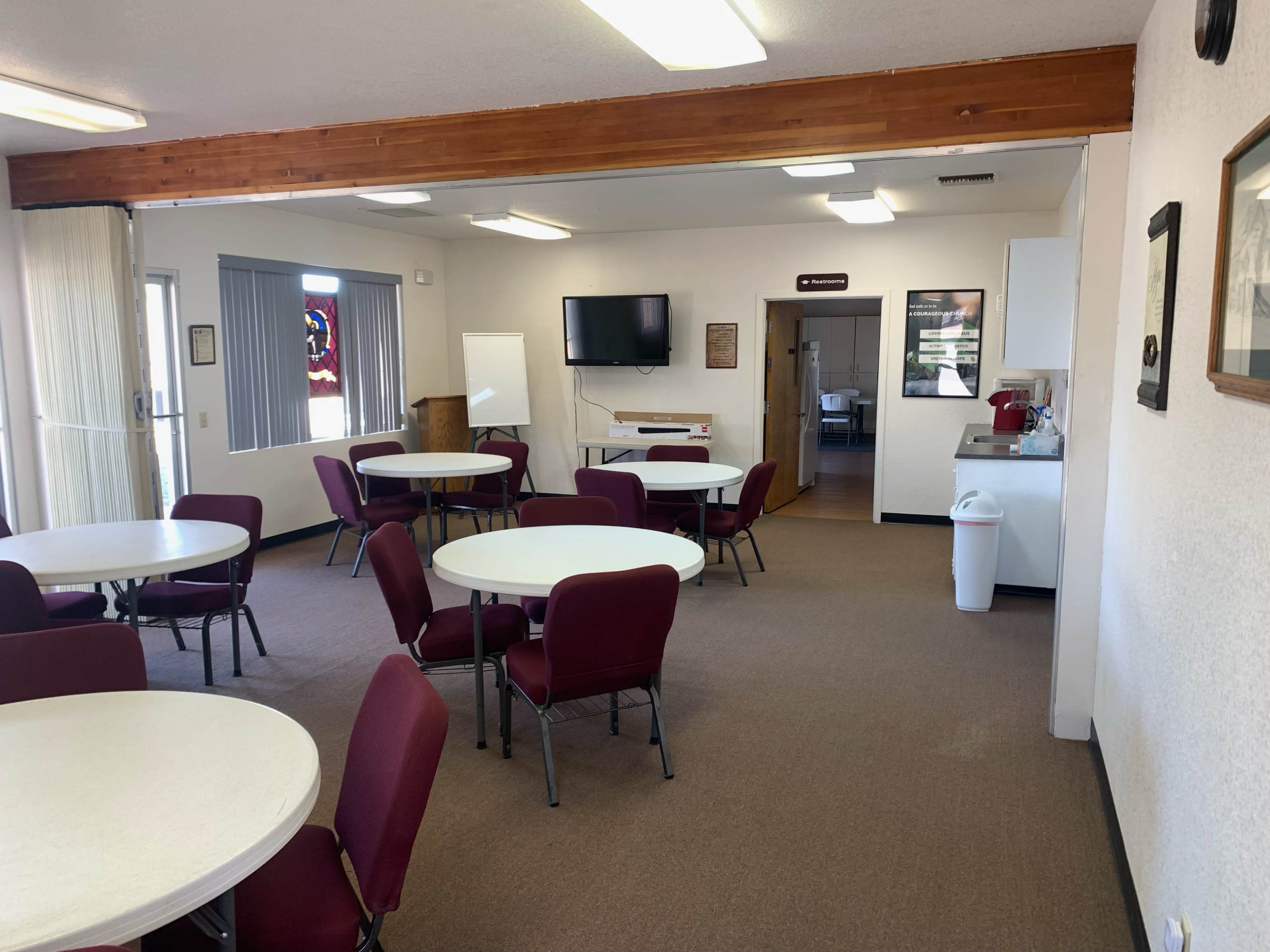 A room with several round tables and maroon chairs, a television mounted on the wall, and a kitchen area in the background.