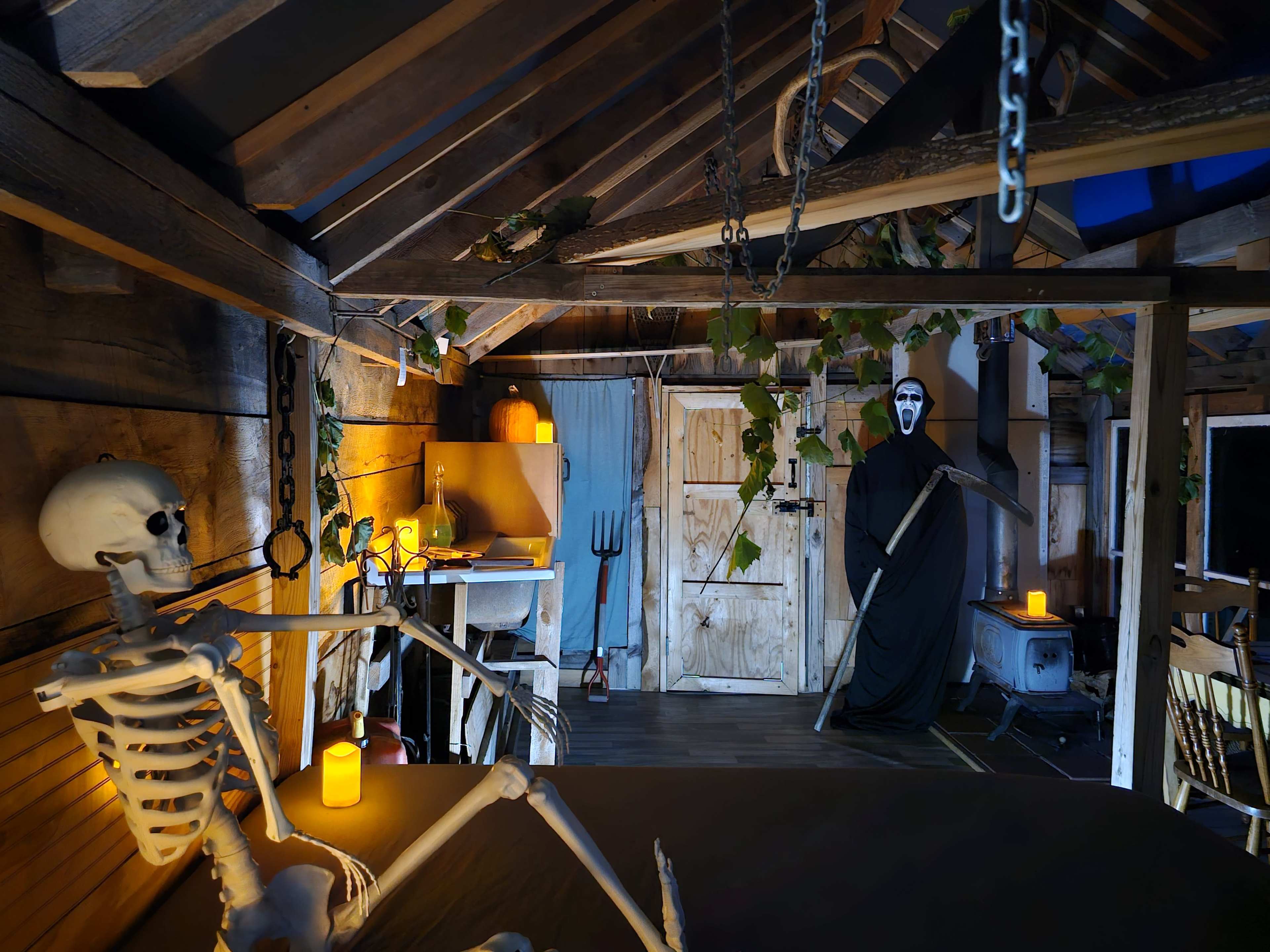 A dimly lit rustic room features a skeleton sitting at a table, while a cloaked figure holding a scythe stands in the corner near wooden doors.