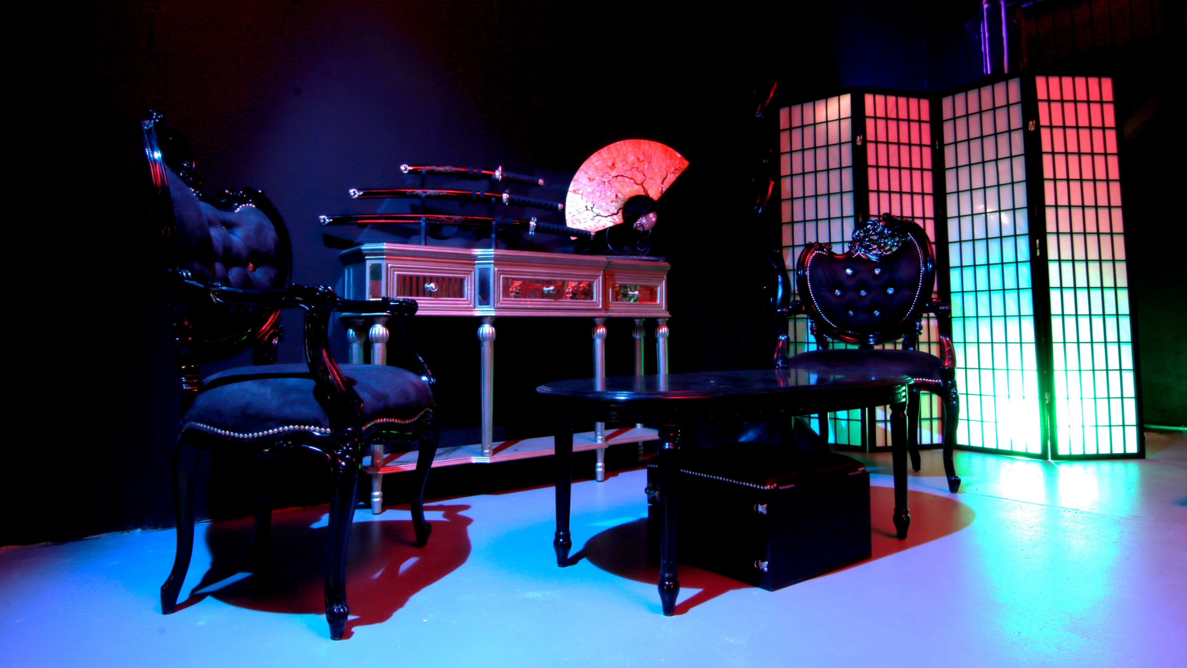 A dark room features two ornate chairs, a low table, a display of decorative swords, and a folding screen illuminated with colored lighting.