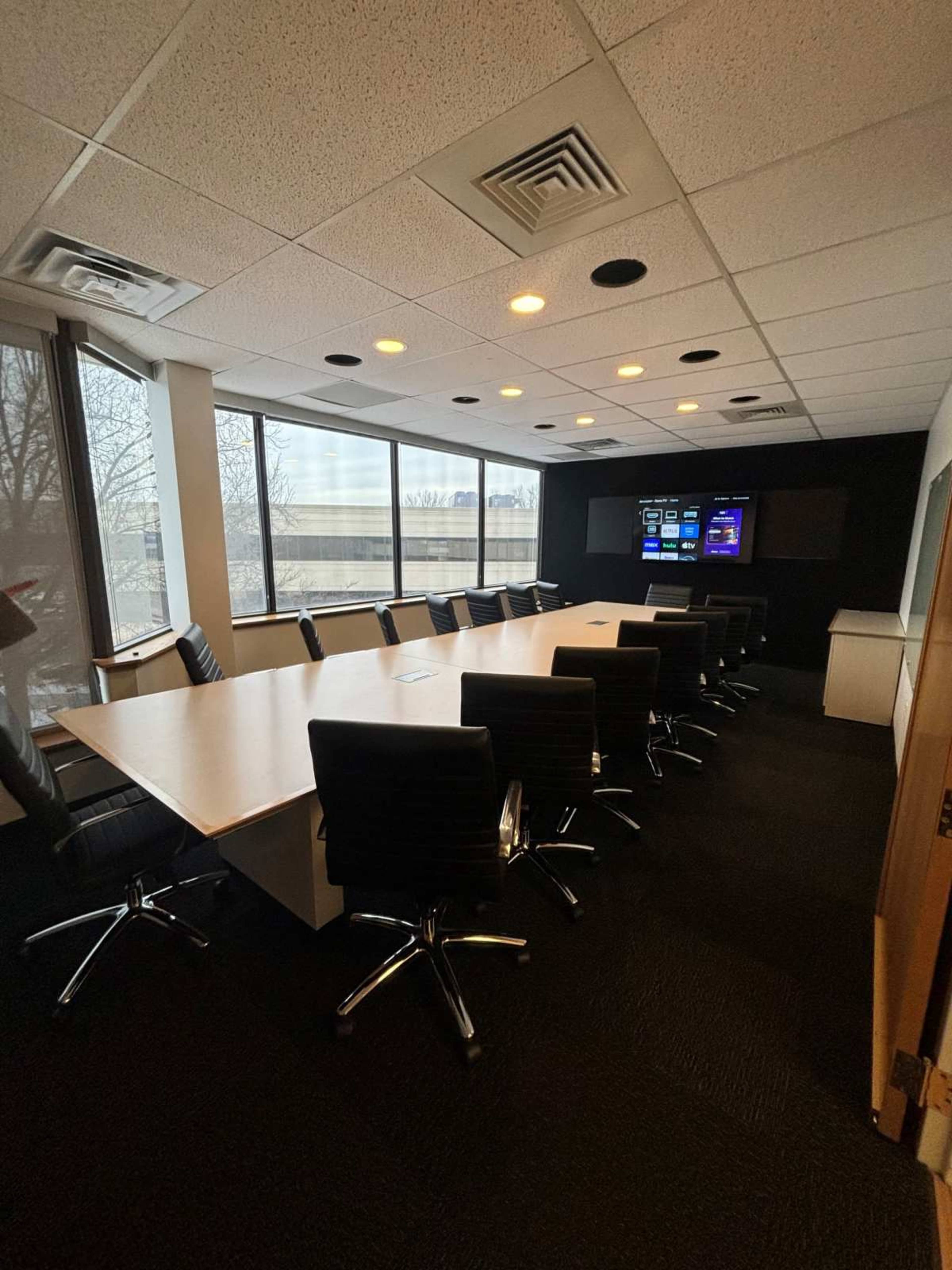 The image shows a modern conference room with a large table, black chairs, and multiple windows overlooking an outdoor area, along with a wall-mounted screen displaying information.