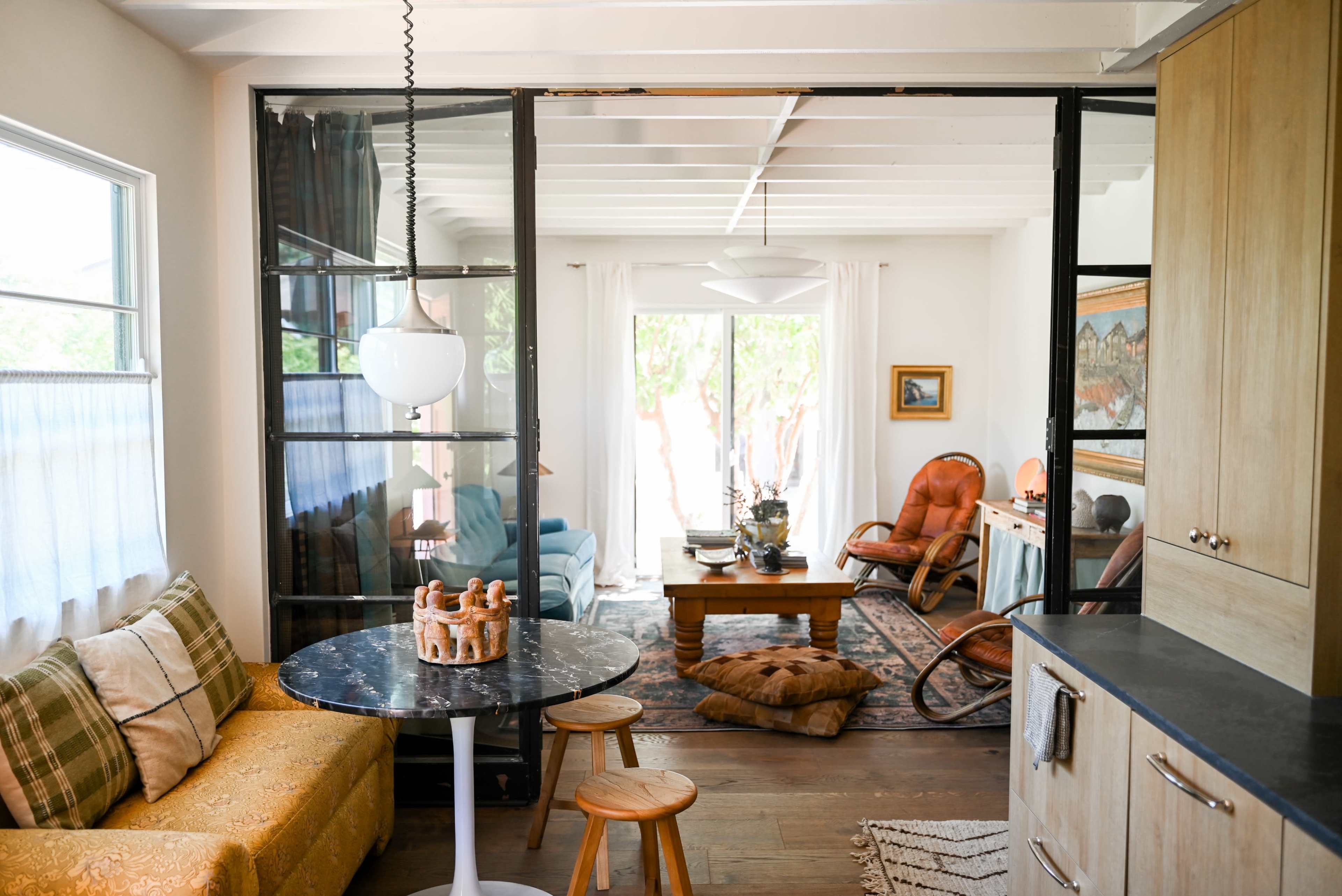 The image shows a bright, open-concept living space featuring a kitchen area with a round table, a cozy seating area with blue and orange furniture, and large windows leading to an outdoor patio.